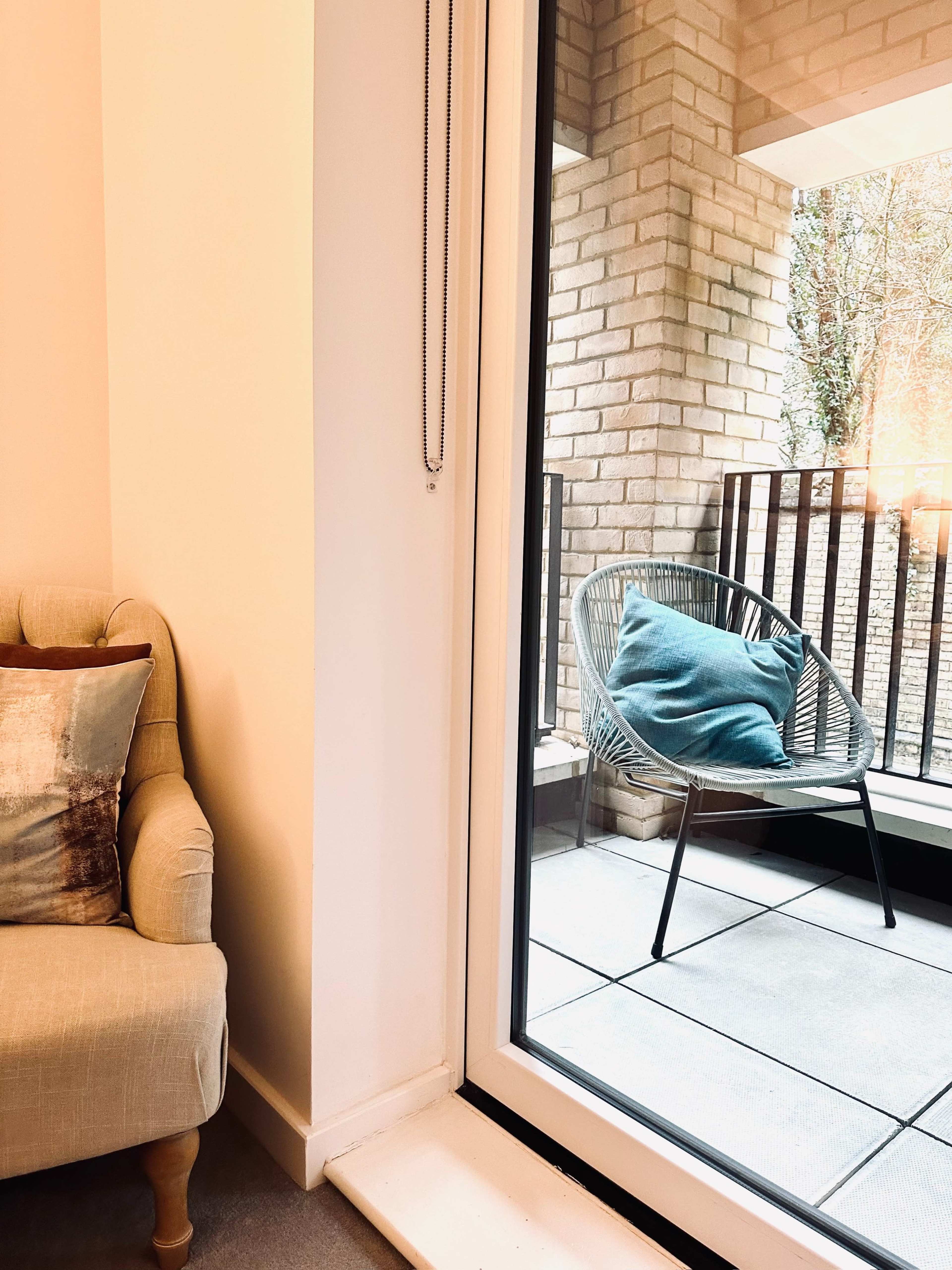 A cozy indoor space with a beige armchair next to a large glass window overlooking a balcony with a single chair and a cushion.