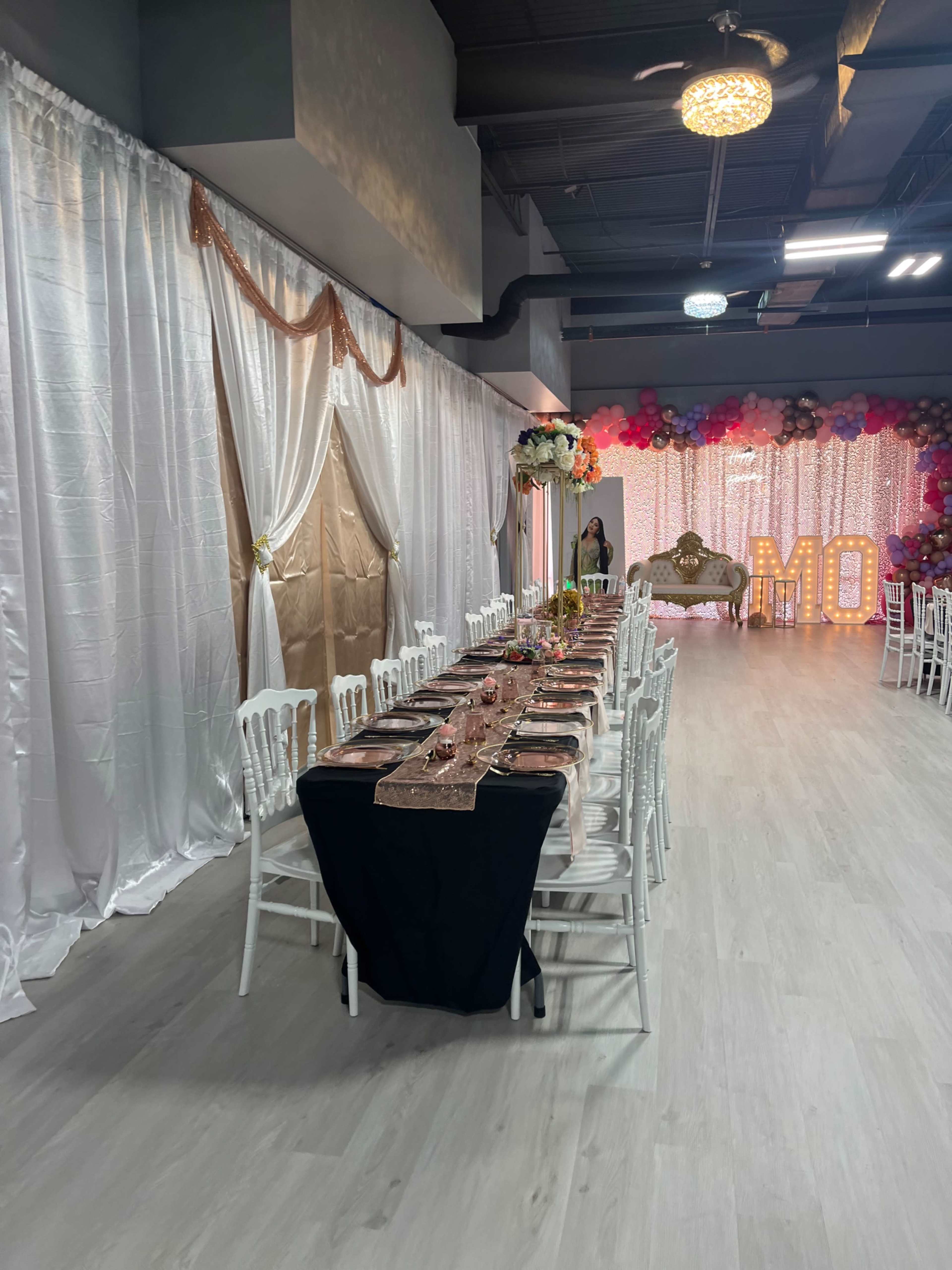 A long dining table is set with elegant tableware and decorations, surrounded by chairs in a banquet hall adorned with shimmering white curtains and colorful balloons.