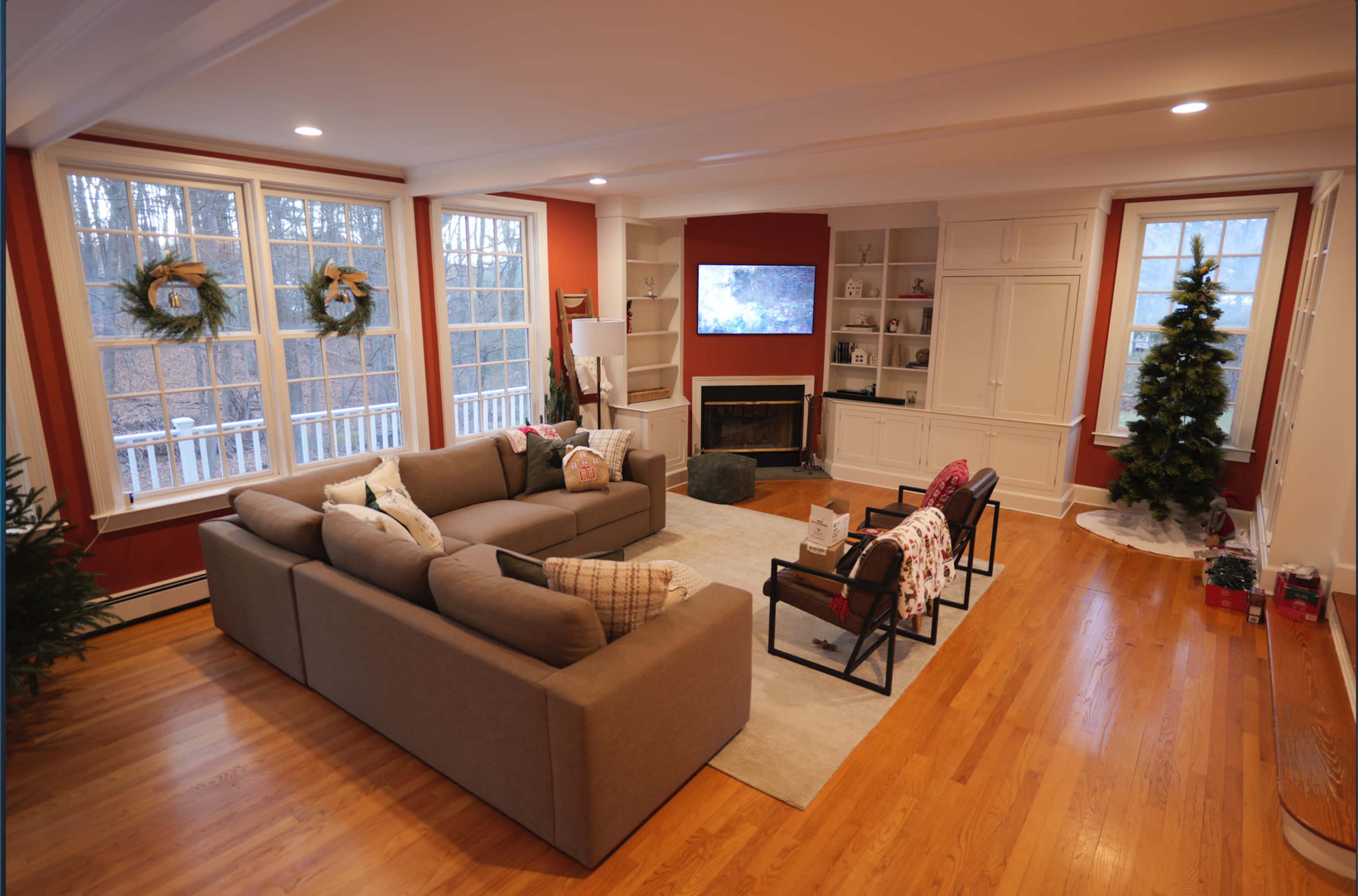 A spacious living room features a sectional sofa, a television mounted on the wall, built-in shelves, and a Christmas tree in one corner.