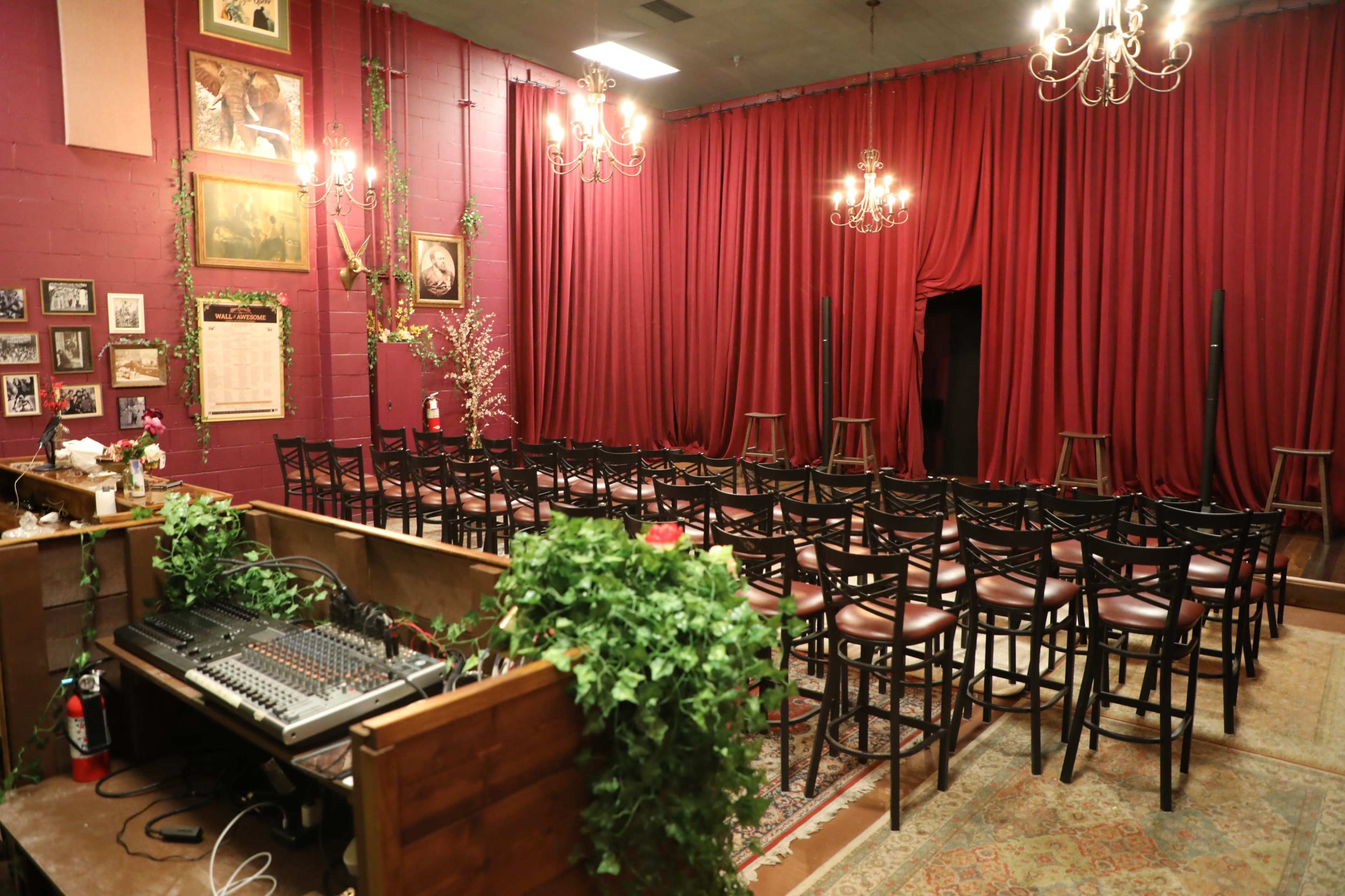 The image shows a small performance space with a red curtain, rows of chairs facing a stage area, and sound equipment positioned to the side.