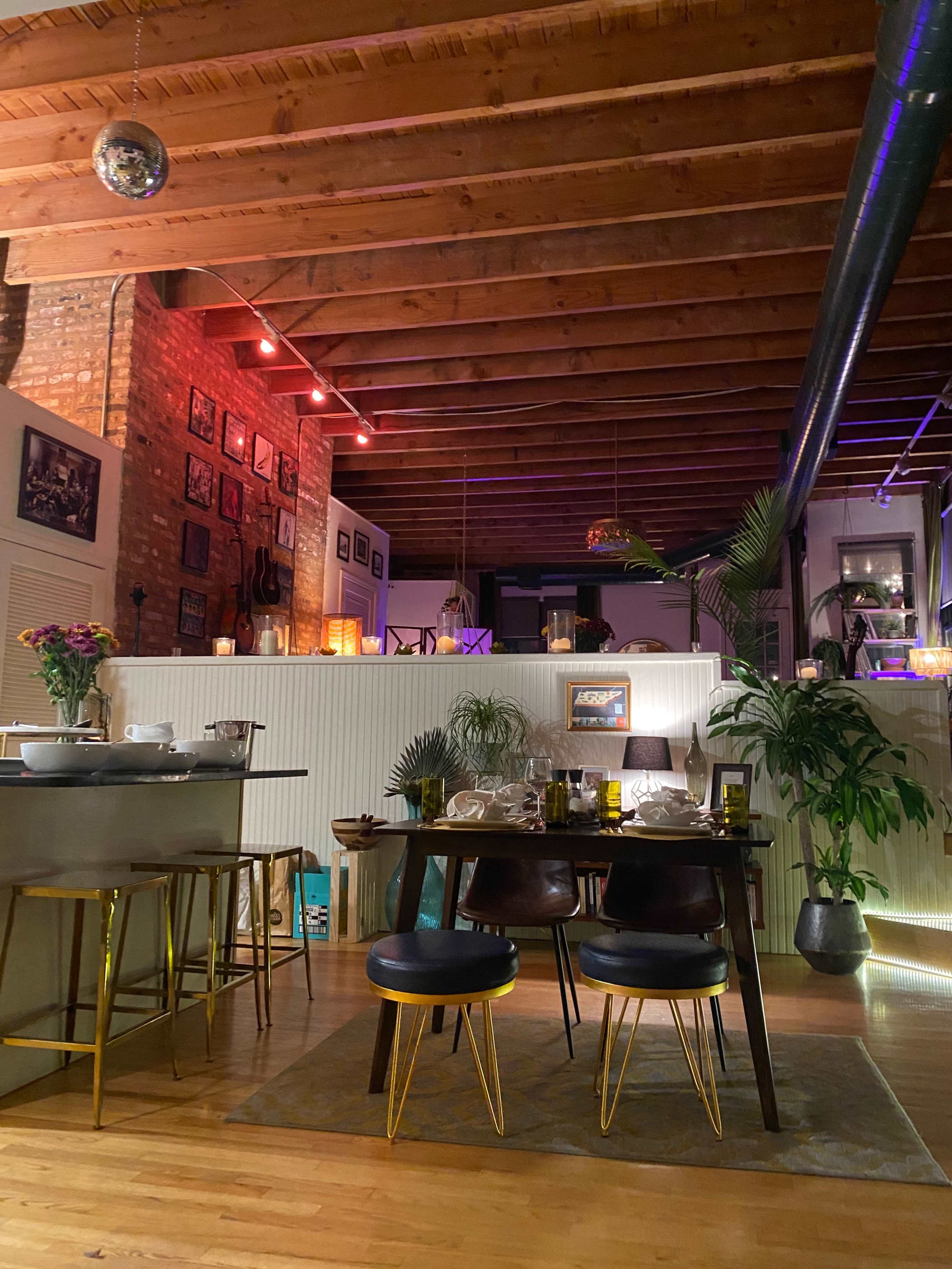 A dining area with a table set for dinner and modern decor is illuminated by colorful lights and features exposed wooden beams above.