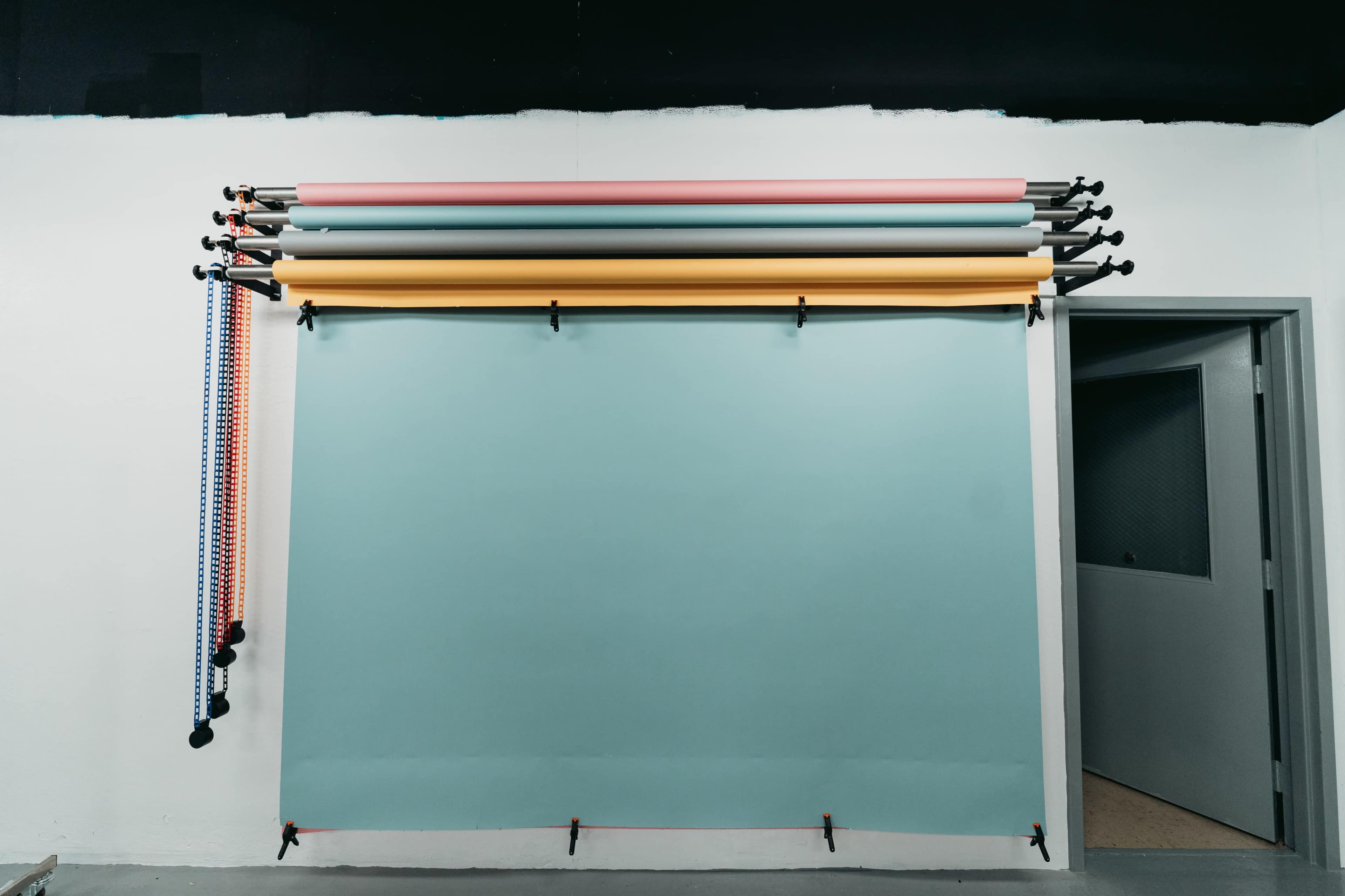 A wall with multiple rolls of colored backdrop paper mounted on a rack, alongside a doorway leading to another room.