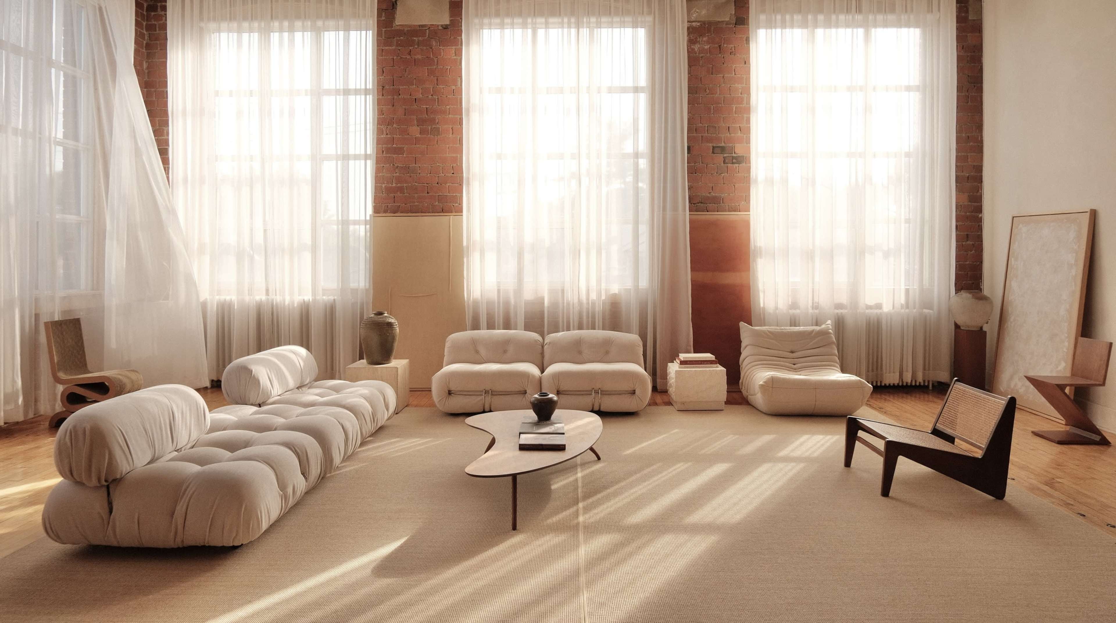 The image depicts a spacious, modern living room featuring a minimalist design with light-colored furniture, large windows allowing natural light, and hardwood flooring.