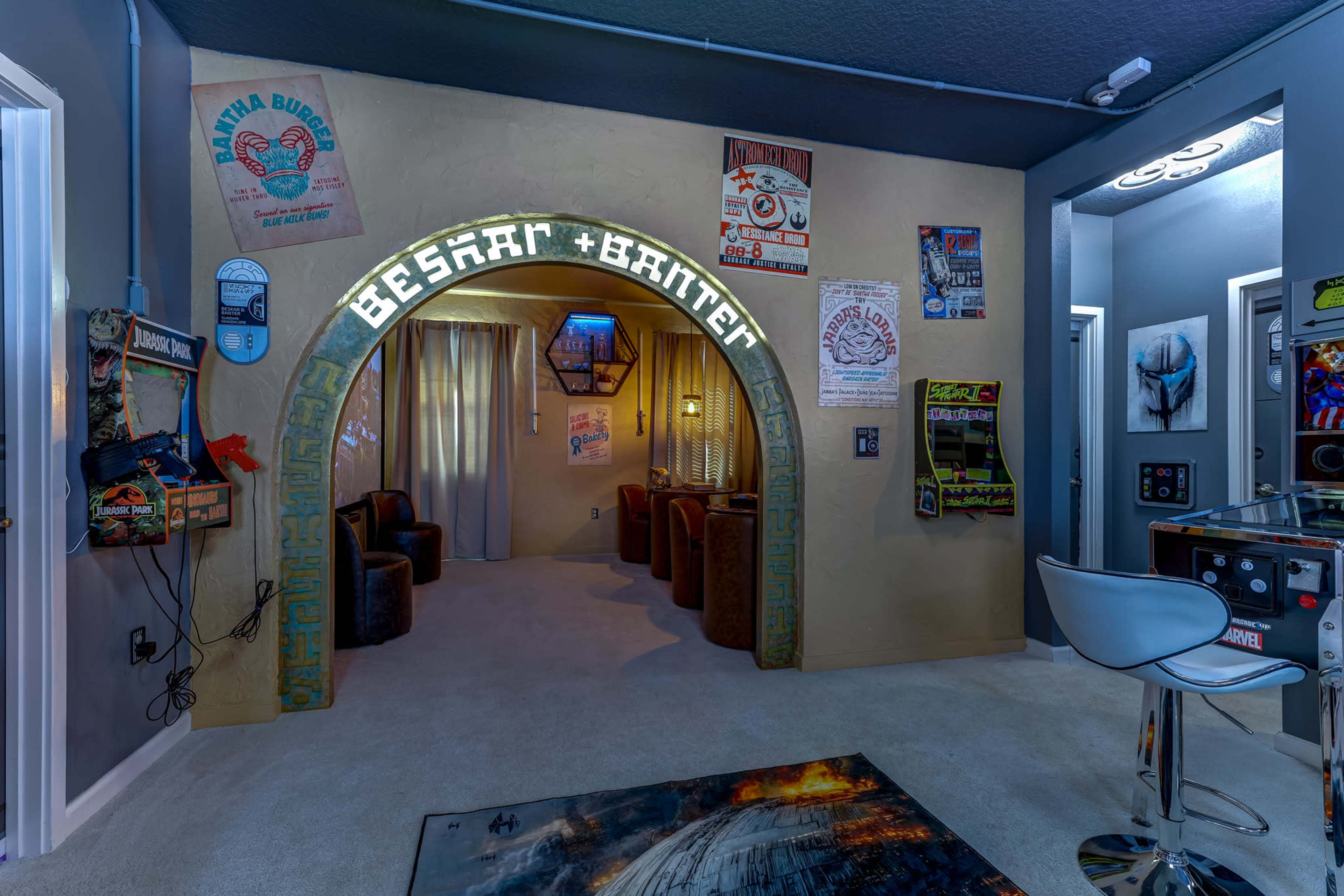 The image shows a retro-style room featuring a curved entrance with the words “BESKAR + BARTER,” arcade machines along the walls, and a seating area with armchairs in a cozy setting.