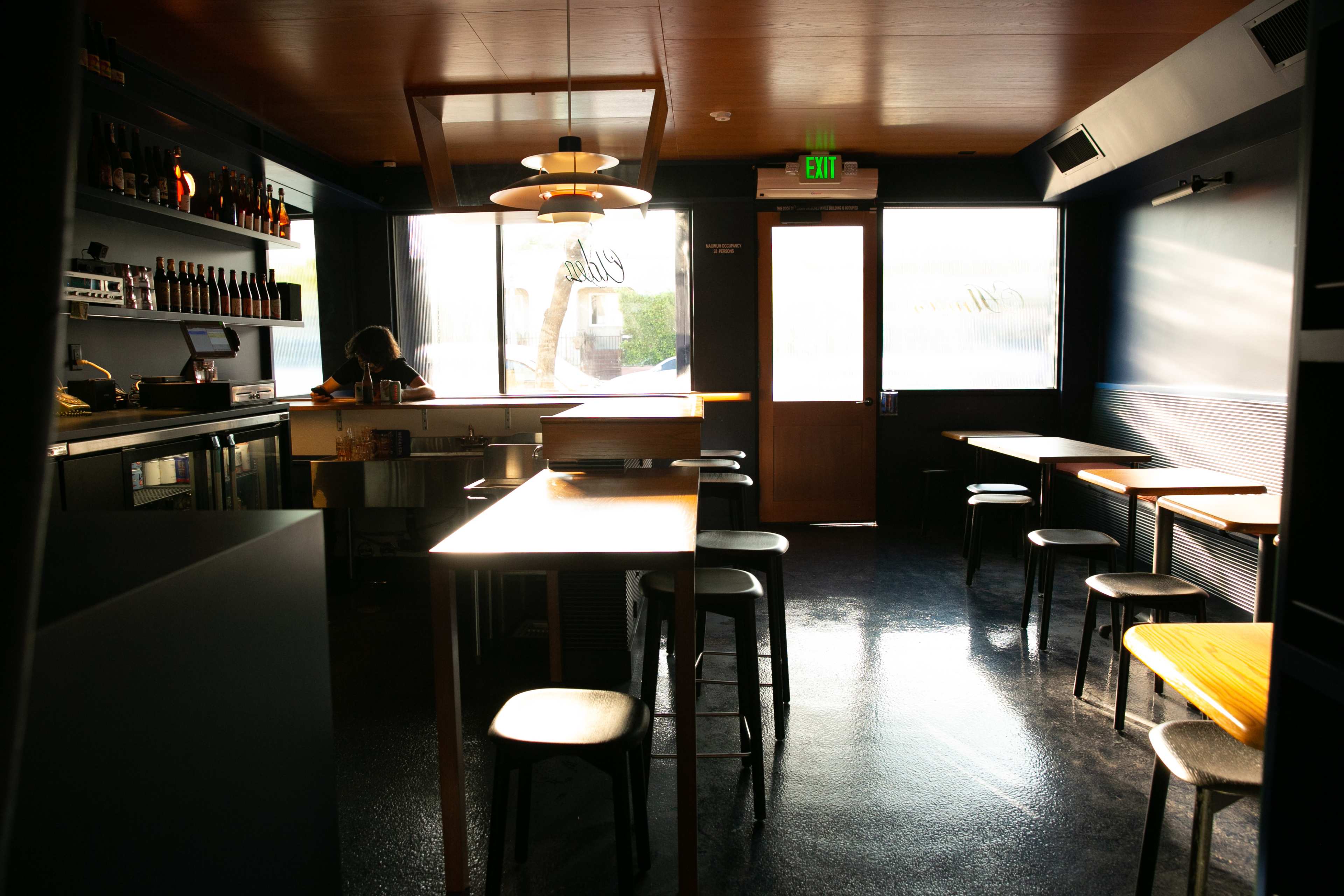 A dimly lit restaurant interior features wooden tables, black stools, a bar area stocked with bottles, and large windows allowing natural light.