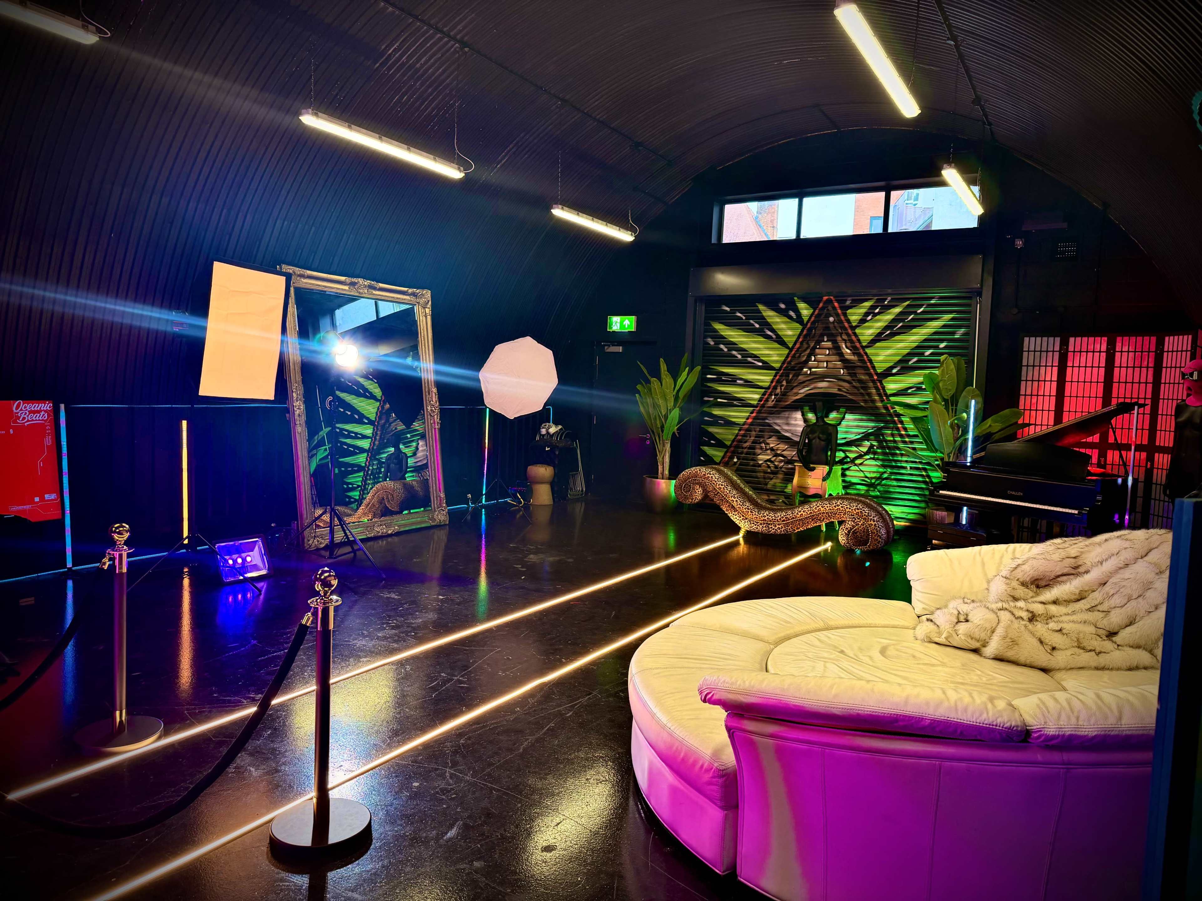 The image shows a well-lit studio space featuring a circular white sofa, a large mirror, tropical plants, and colorful lighting against a backdrop of a vibrant wall mural.