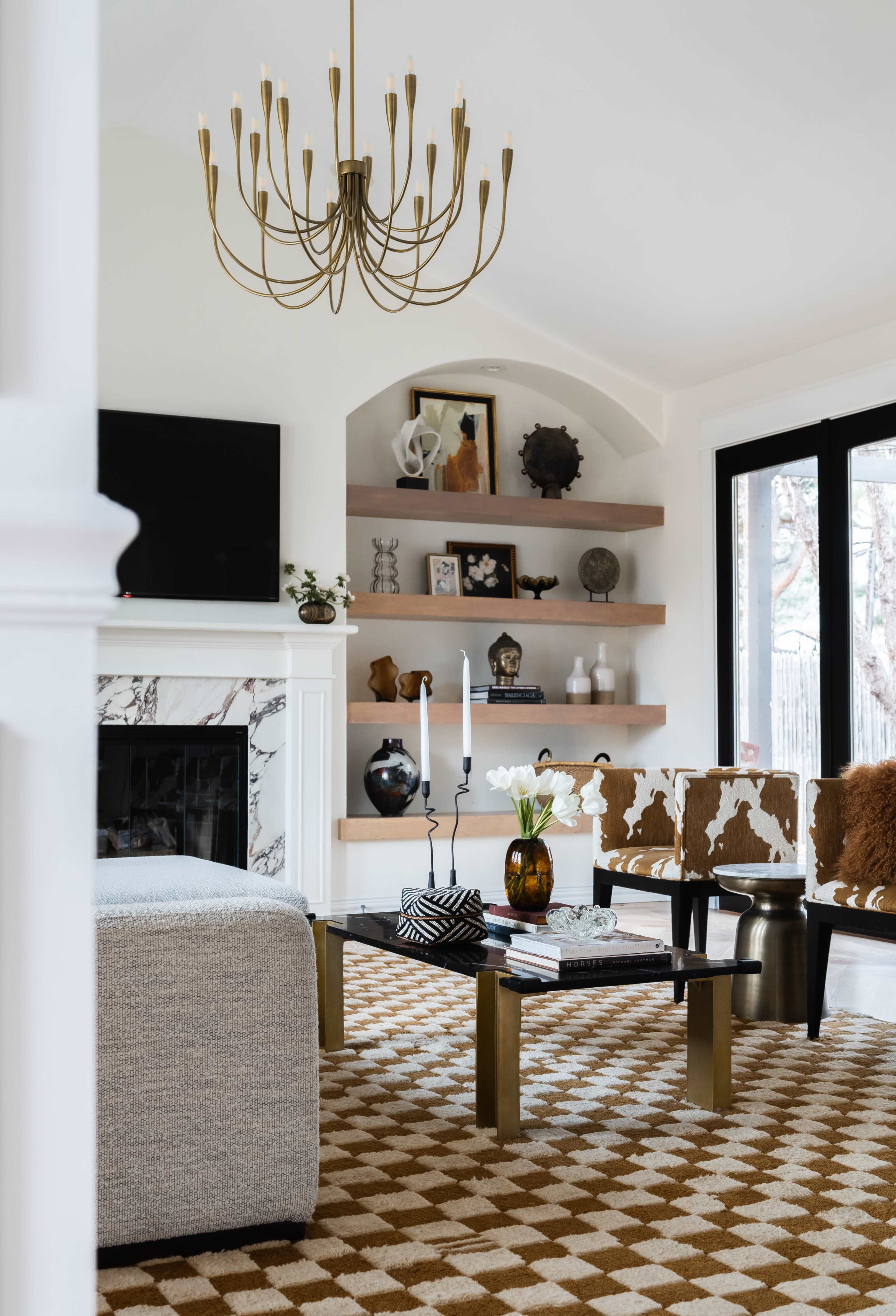 A modern living room features a chandelier, a marble fireplace, shelves with decor, a patterned rug, and stylish furniture.