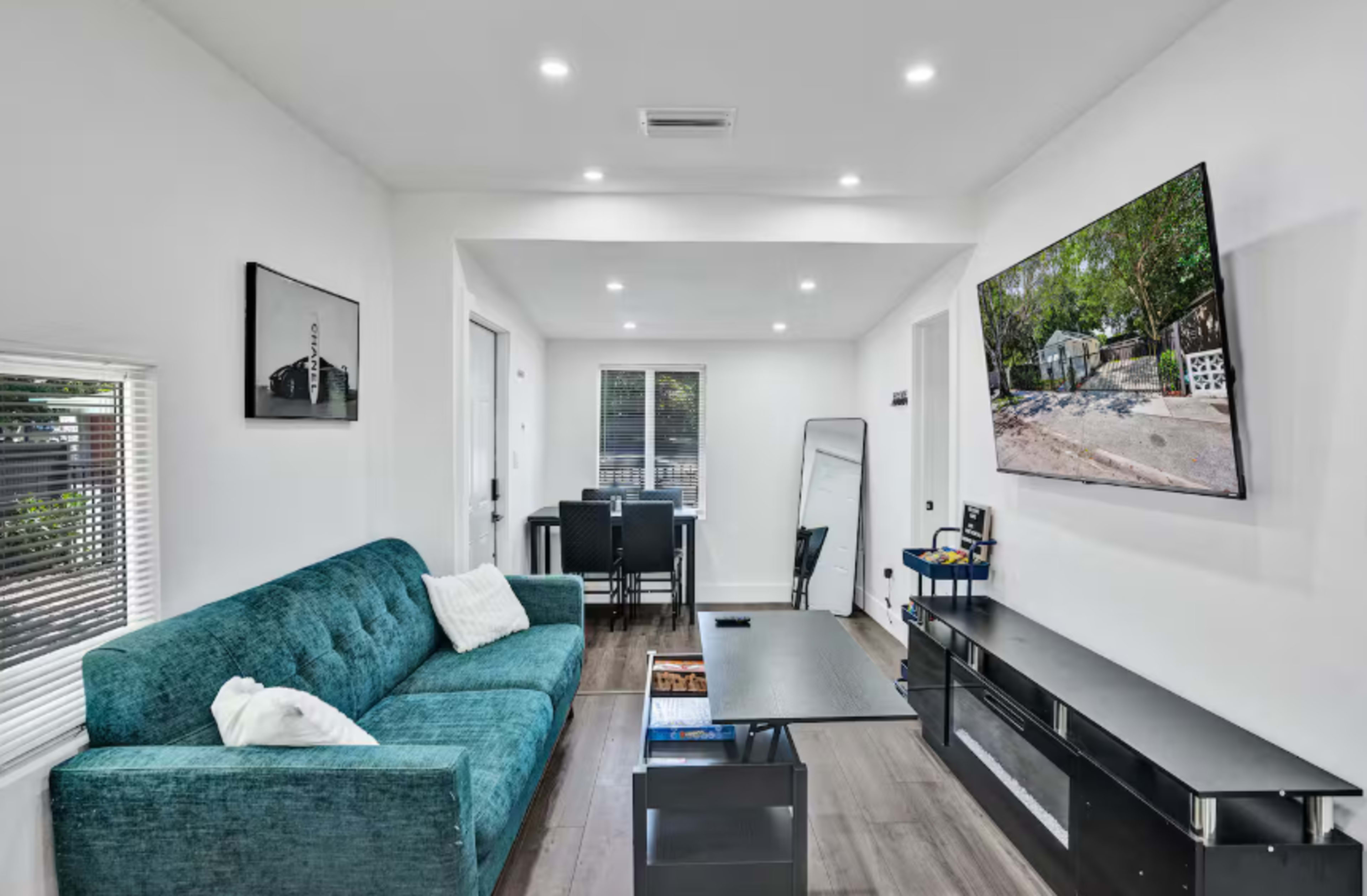 The image shows a modern living room featuring a teal couch, a black coffee table, a wall-mounted television, and a dining area in the background.