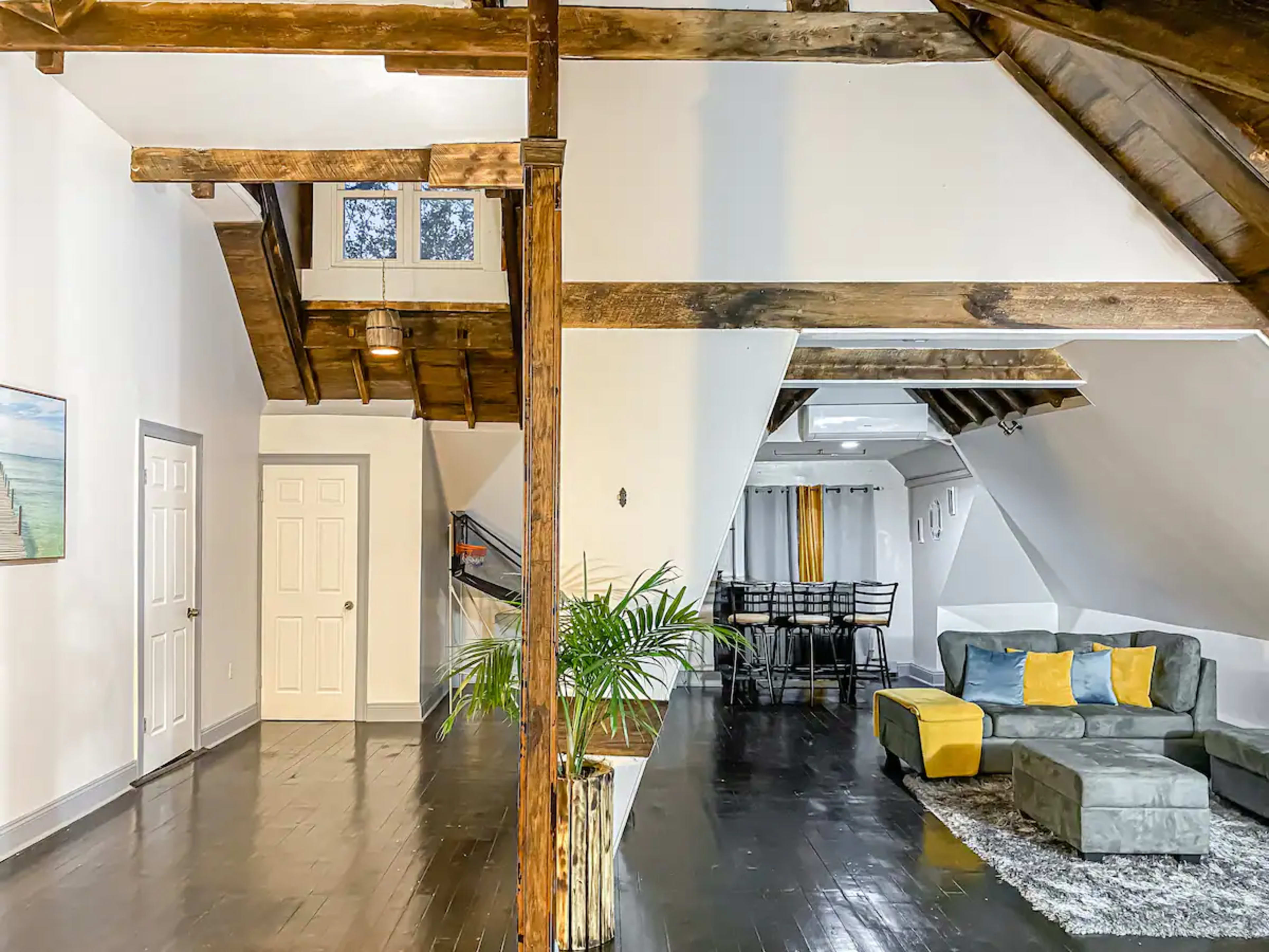A spacious interior with exposed wooden beams, a staircase leading to an upper level, and a living area featuring a gray couch with yellow and blue cushions.