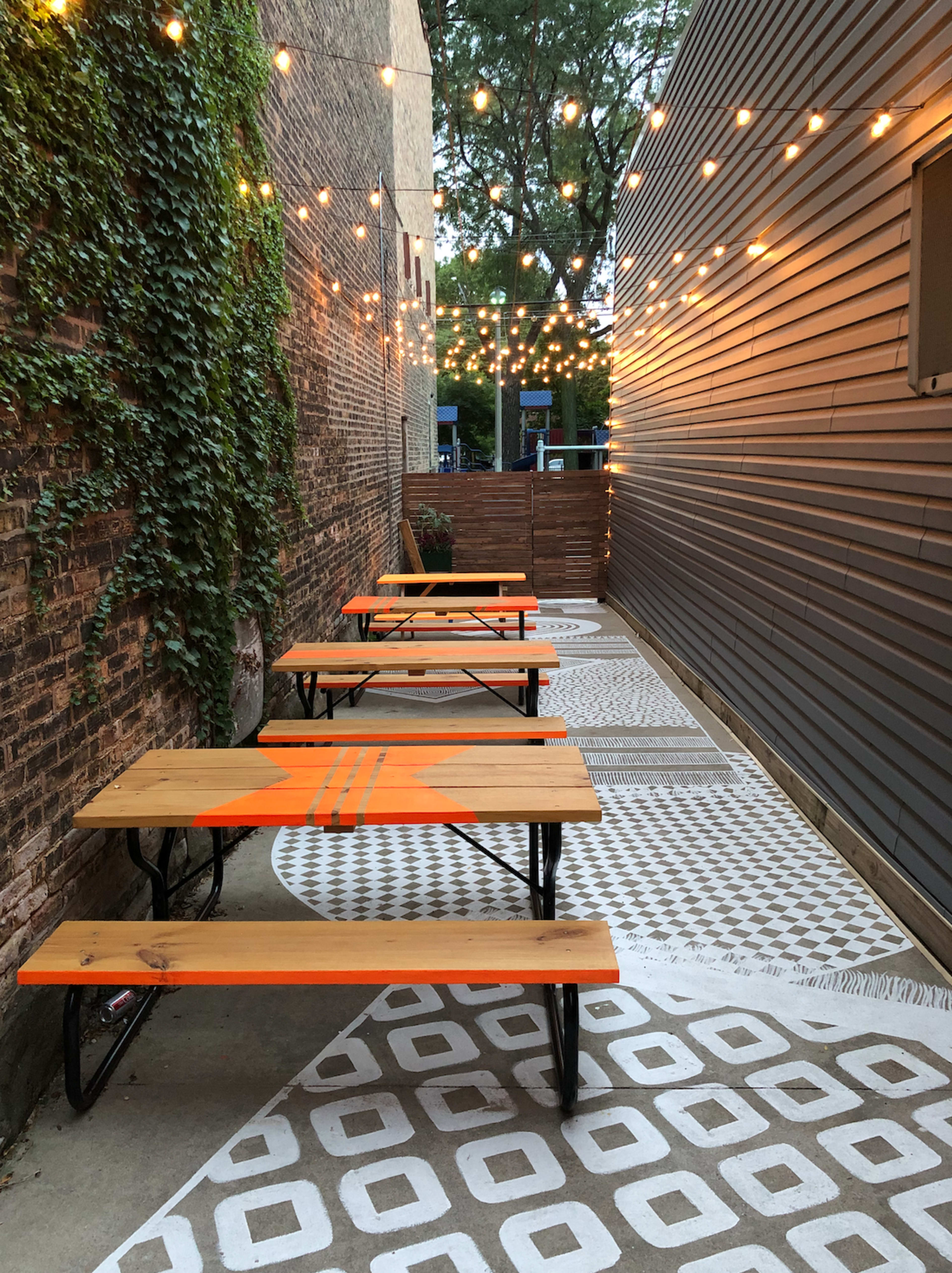 A narrow outdoor space features several wooden tables with orange accents, string lights overhead, and patterned concrete flooring.