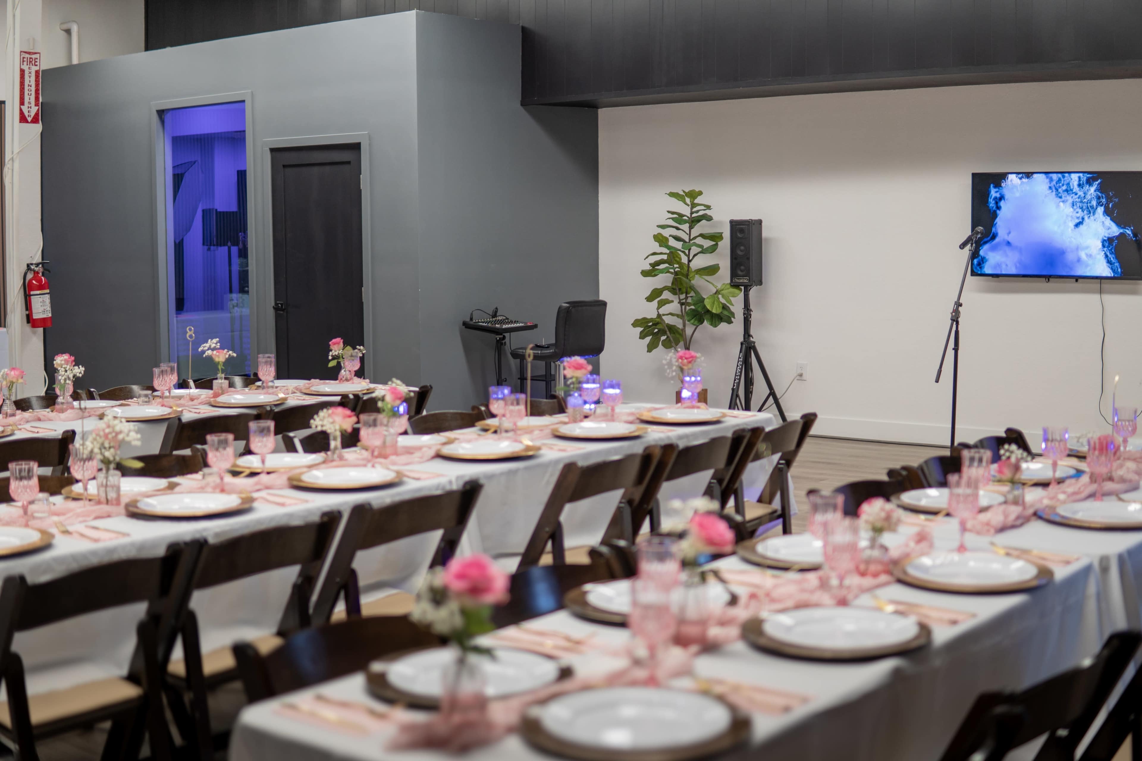 The image shows a rectangular banquet setup with neatly arranged tables, adorned with pink floral centerpieces and set for a meal, in a well-lit event space featuring a microphone and a speaker nearby.