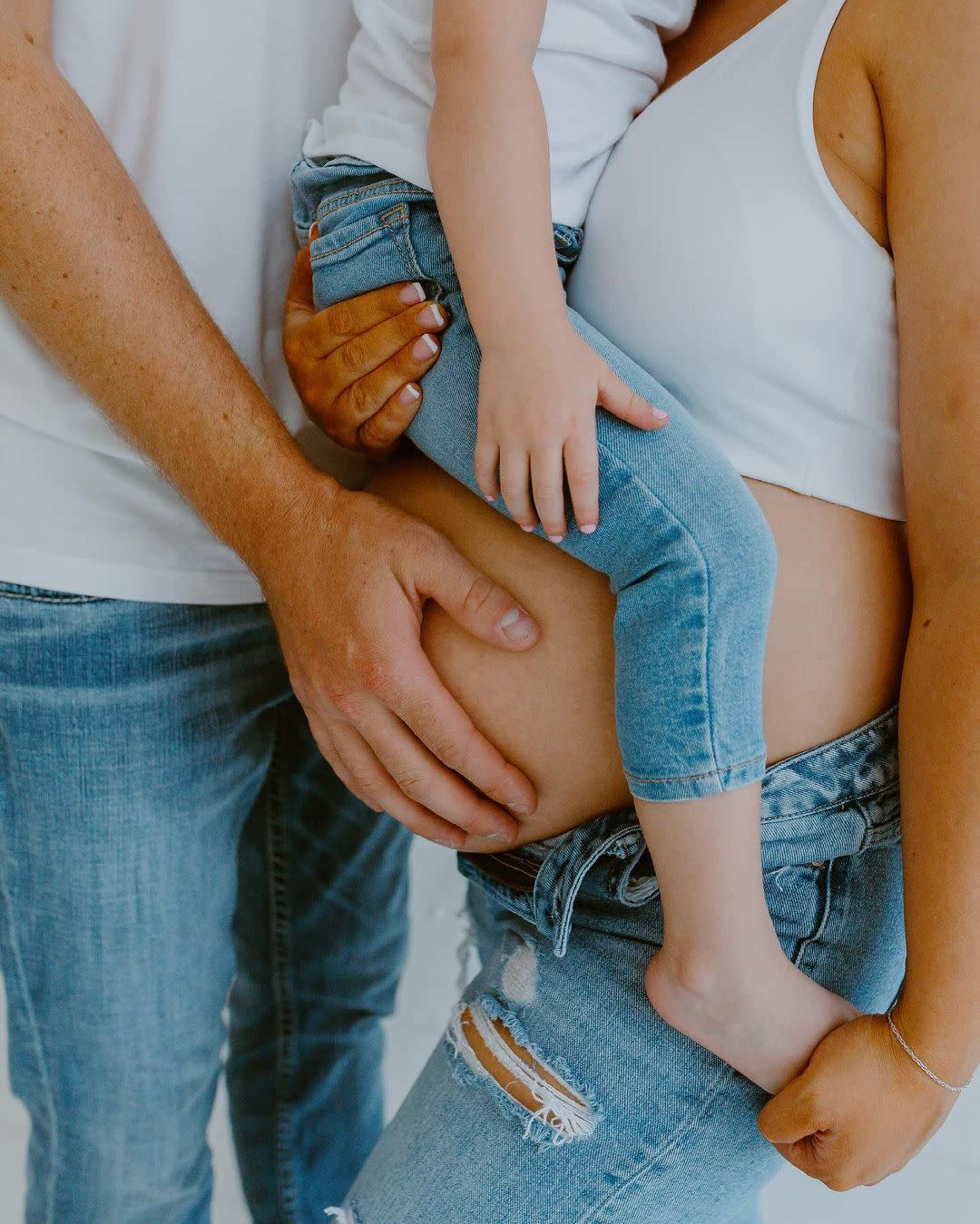 A pregnant woman stands with her partner, who gently holds her belly, while a young child sits on her hip.