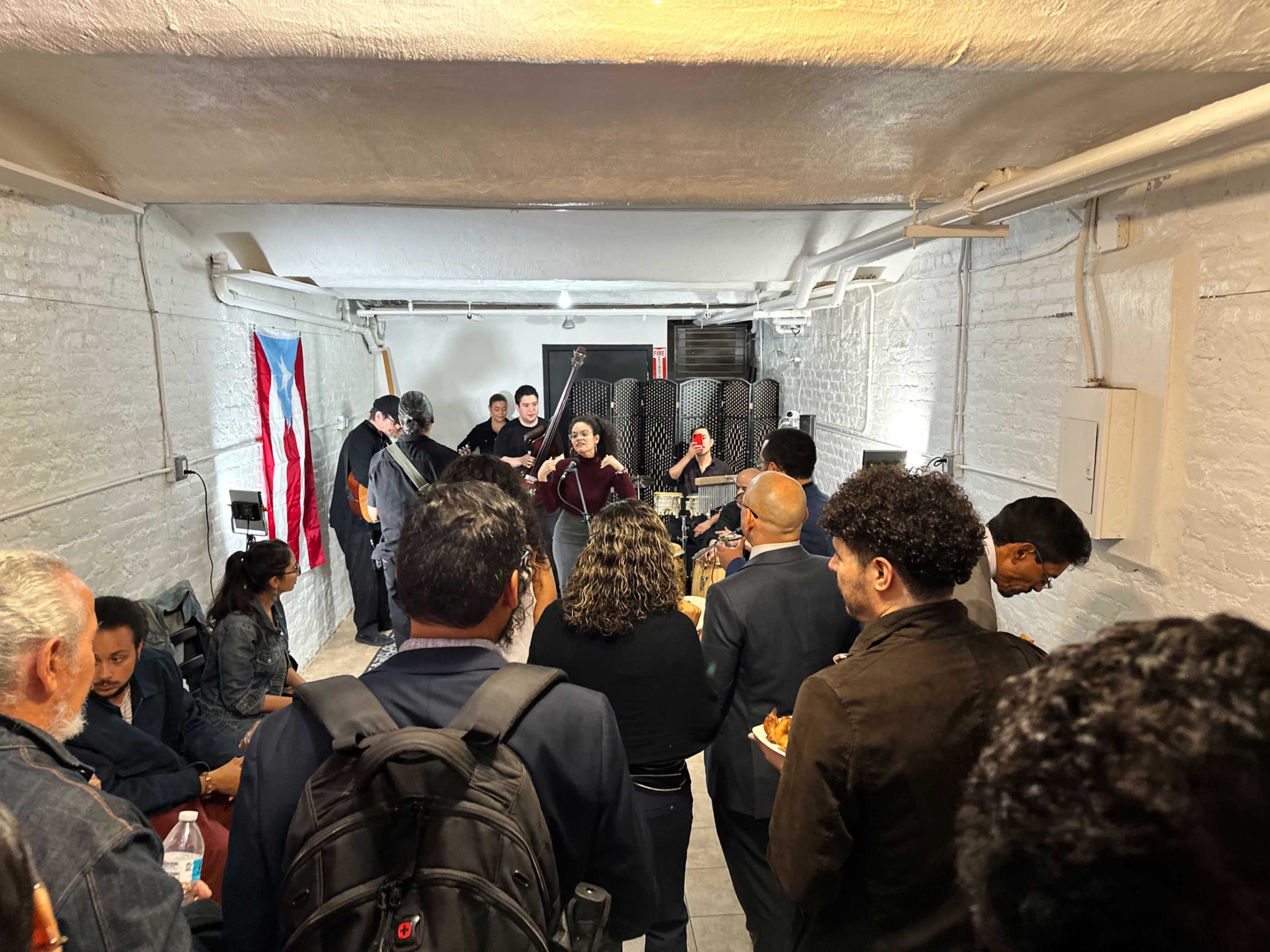 A group of people gathers in a narrow, white-walled room with a musician performing in the background.
