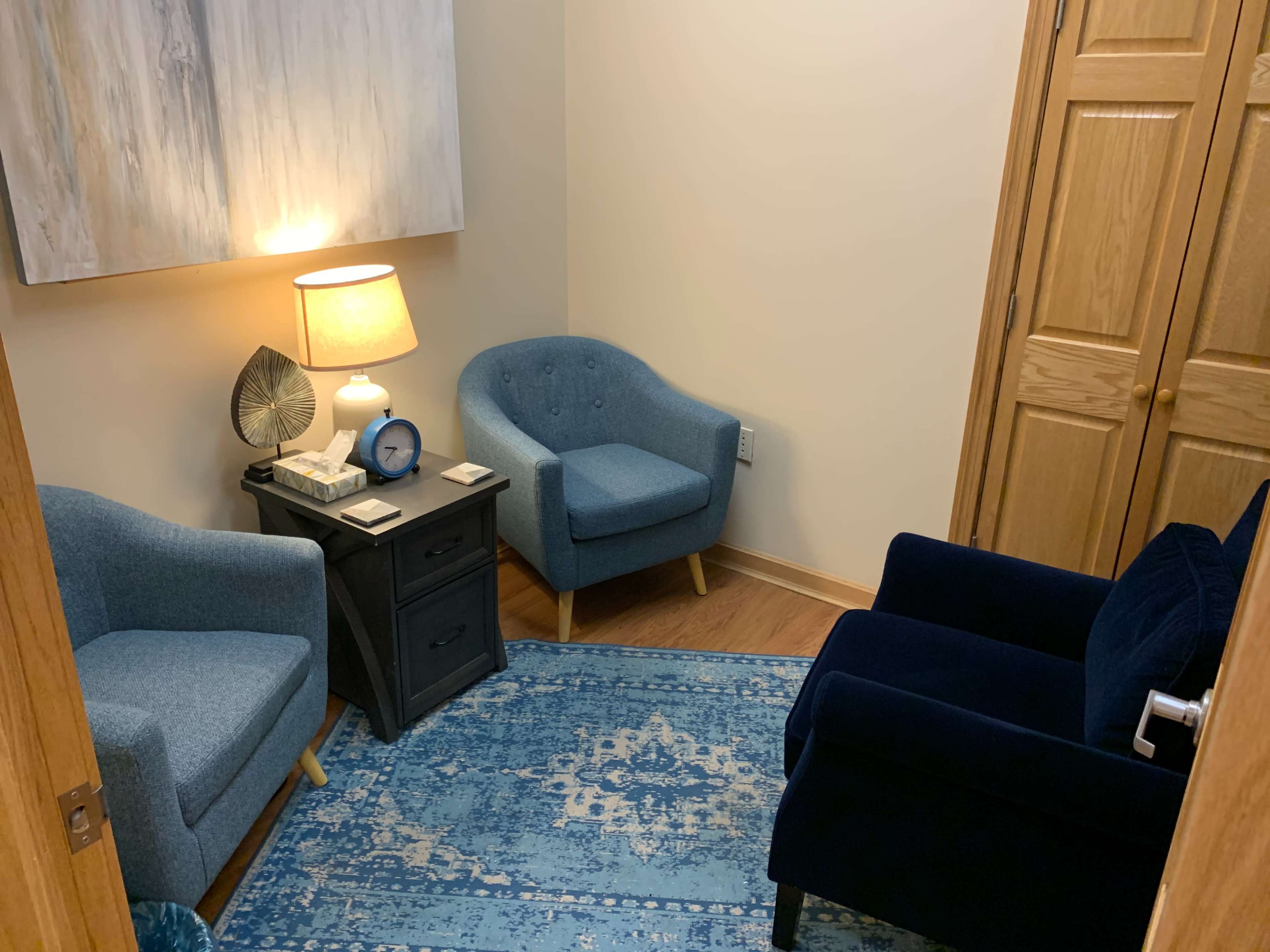 The image shows a small, softly lit room featuring two blue chairs, a dark nightstand with a lamp and clock, and a light blue area rug on a wooden floor.