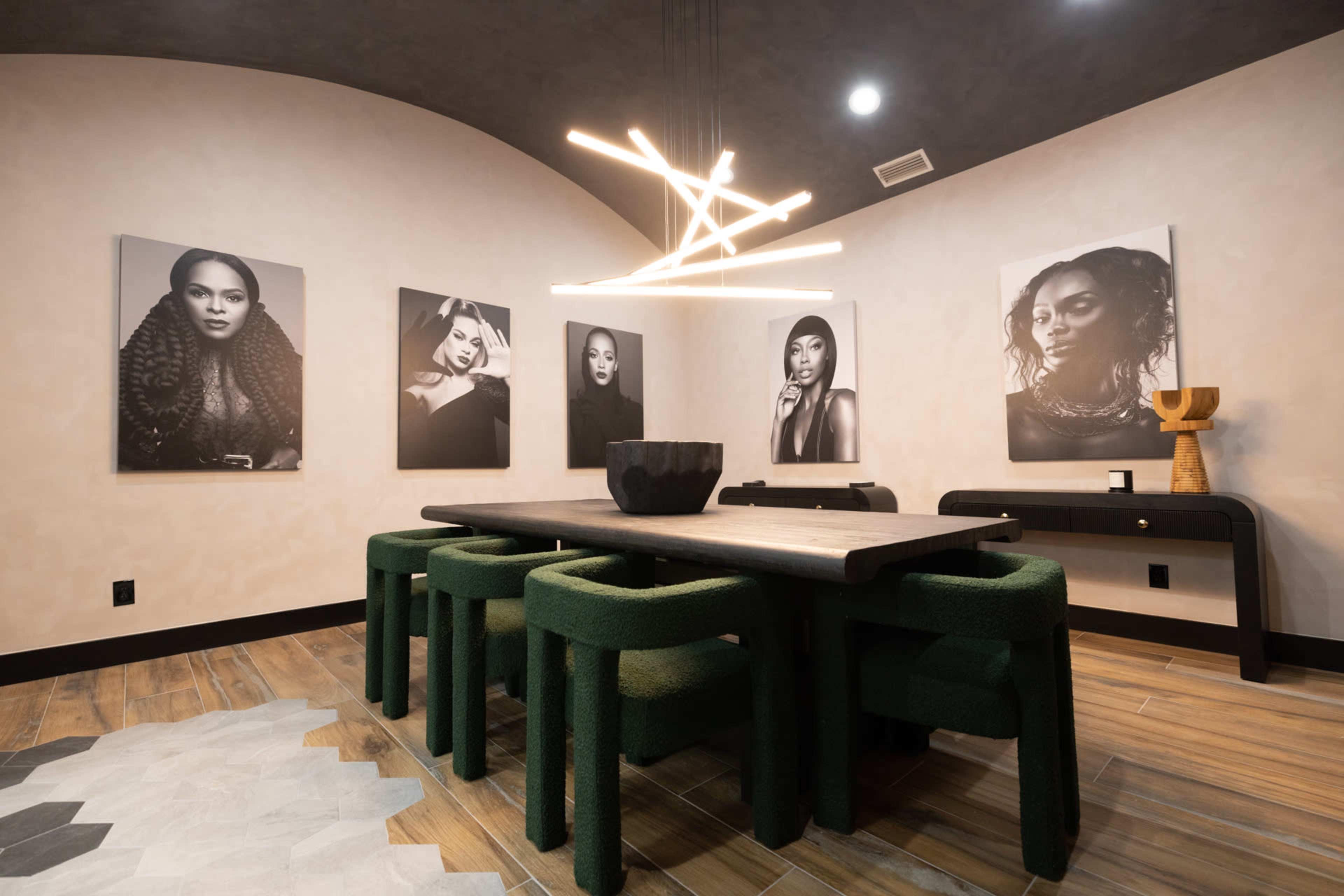 The image depicts a modern dining room featuring a wooden table surrounded by green fabric chairs, with black and white portrait photographs on the walls and a contemporary light fixture overhead.