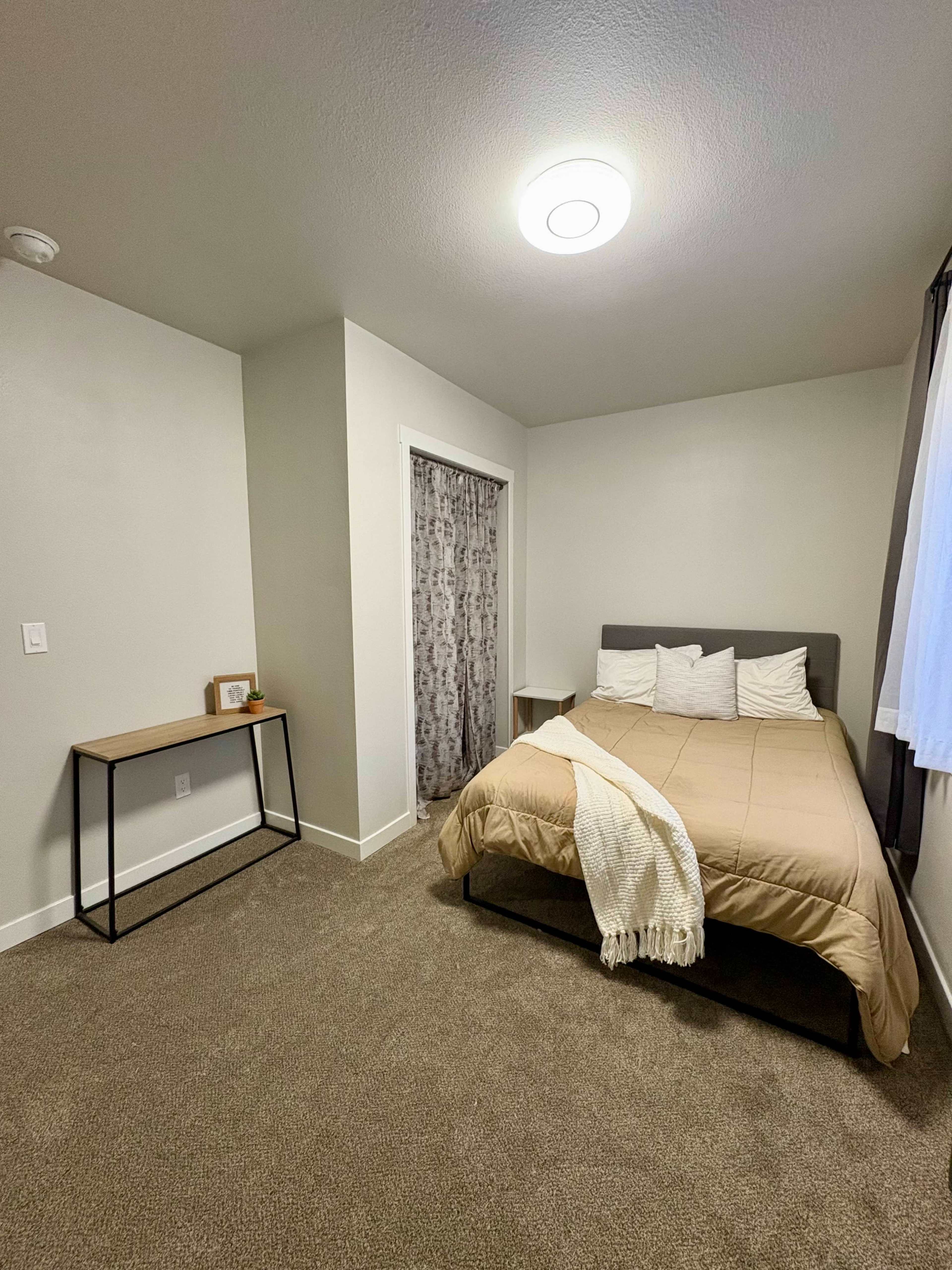 The image shows a neatly arranged bedroom featuring a bed with beige bedding, accompanied by a small table and a closet with a curtain.