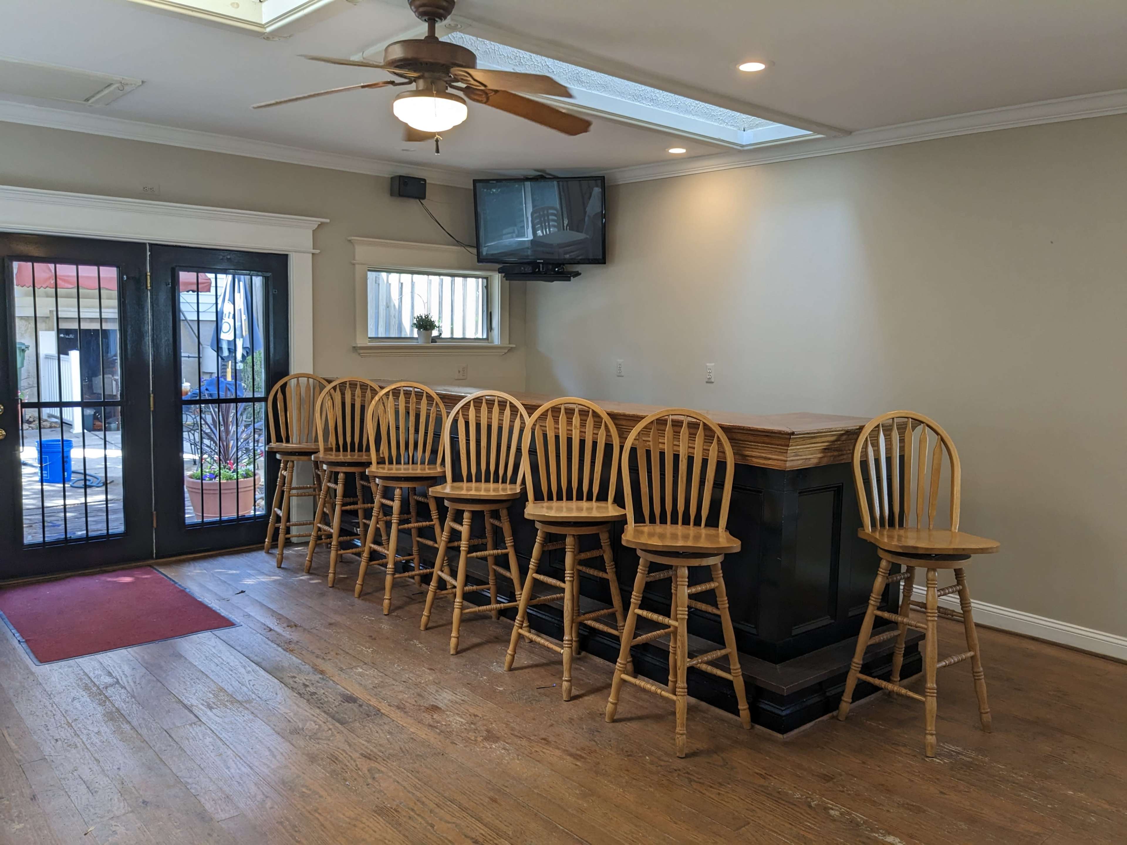 A wooden bar with eight tall, seated chairs facing a wall-mounted television, situated in a room with large glass doors leading to an outdoor area.