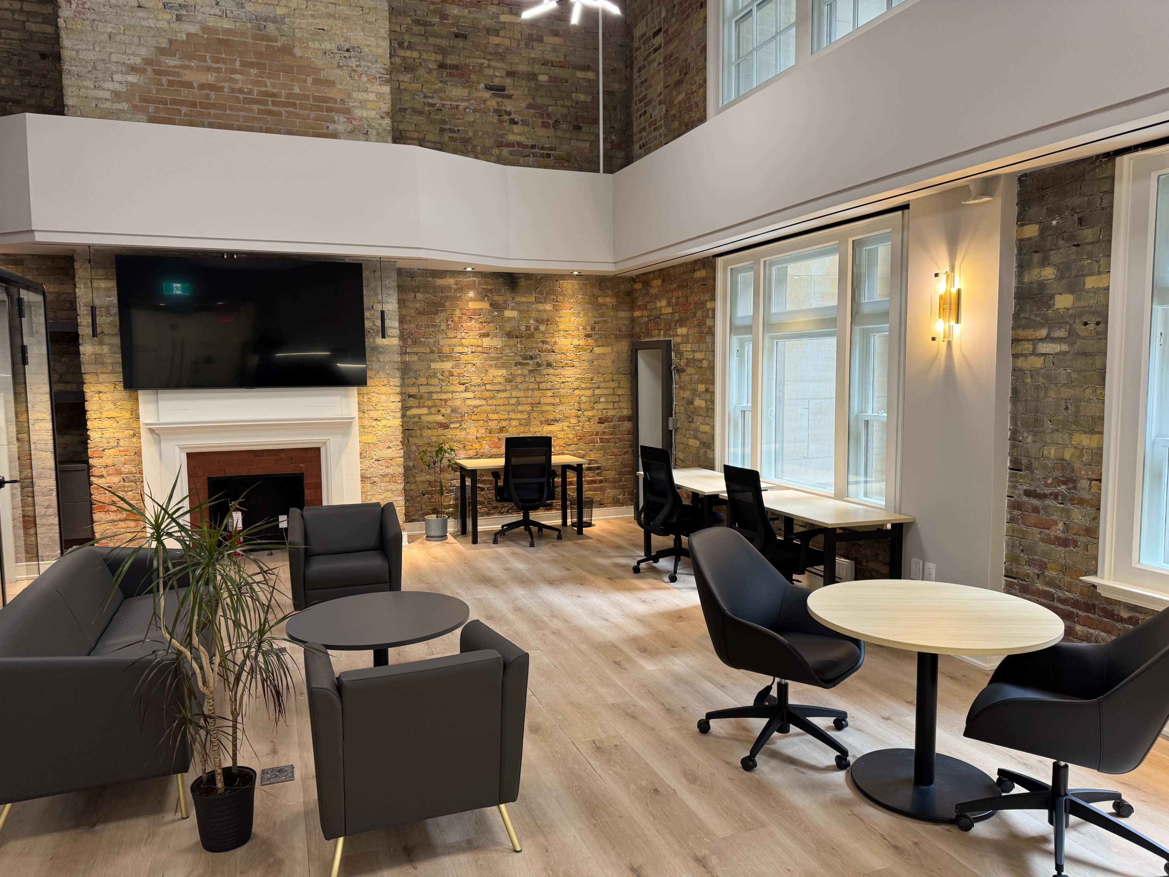 The image shows a modern office space featuring a combination of seating areas and workstations, with exposed brick walls and large windows providing natural light.