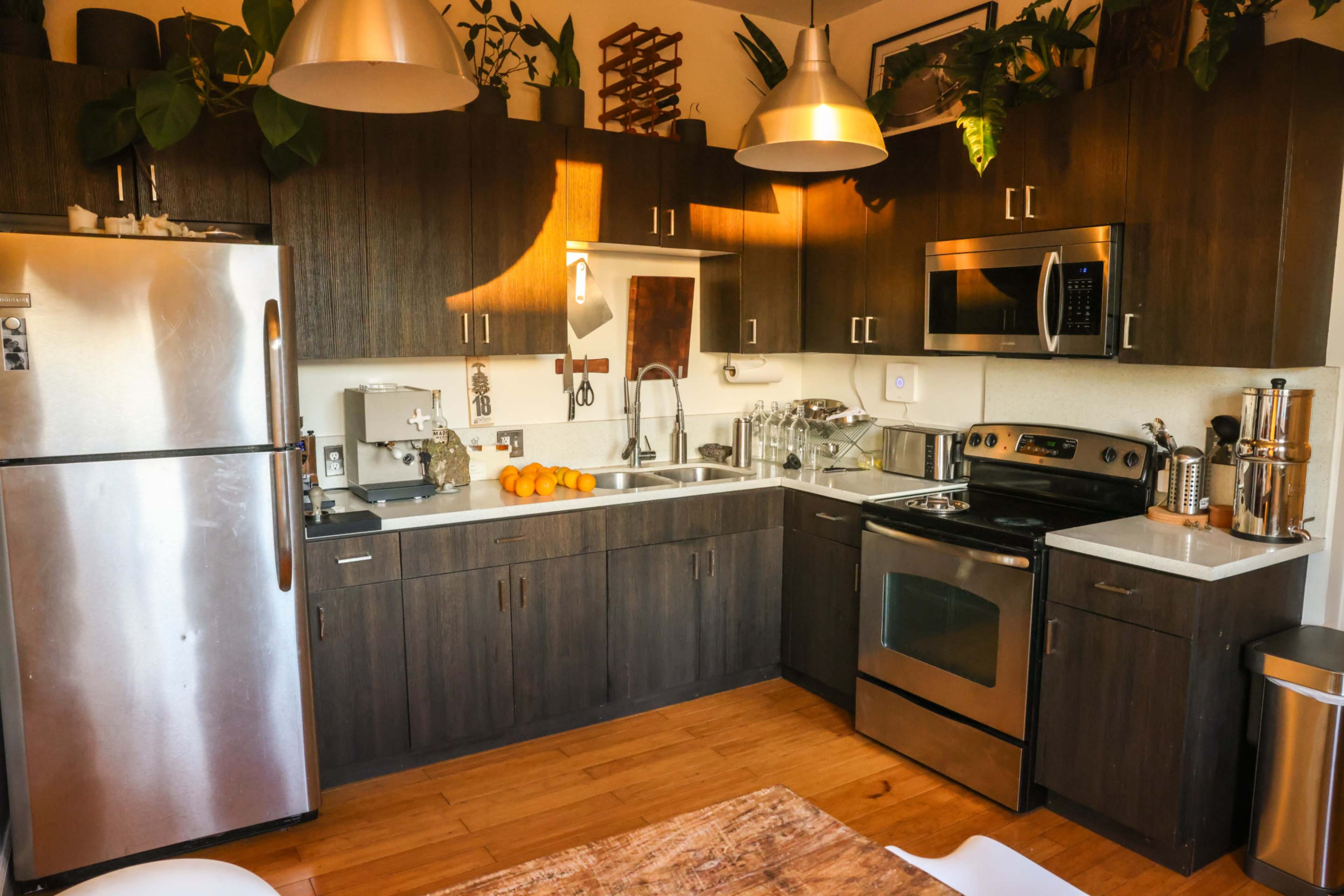 The image shows a modern kitchen with dark wooden cabinetry, stainless steel appliances, and a countertop featuring several utensils and orange fruits.