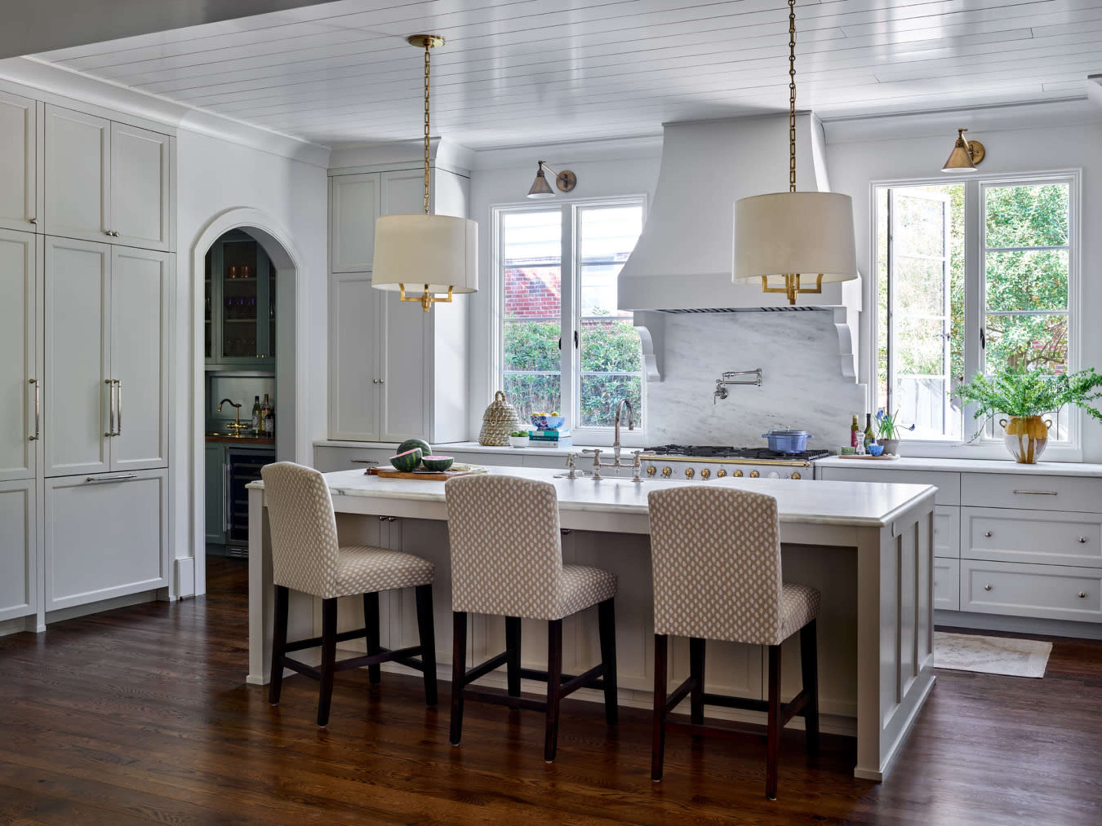 A modern kitchen features a central island with bar stools, white cabinetry, and large windows letting in natural light.