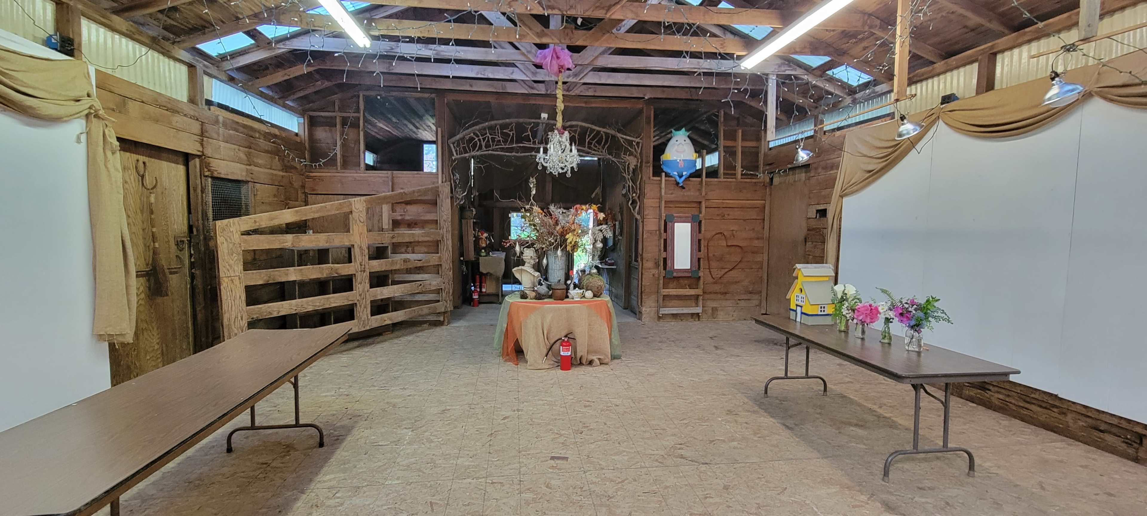 The interior of a rustic barn features a central table with floral arrangements, flanked by two long tables and wooden walls adorned with drapery.