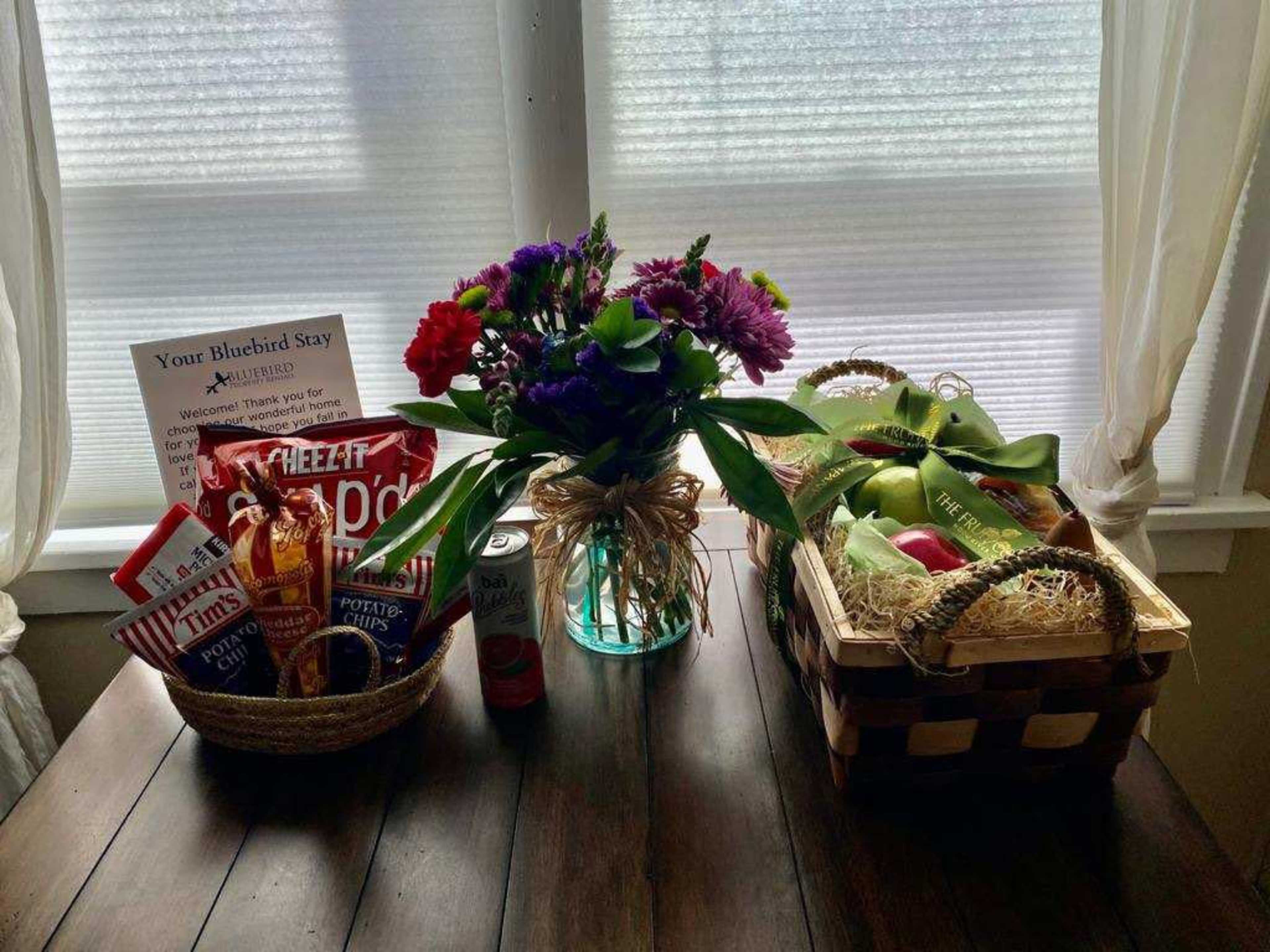 The image shows a wooden table with two wicker baskets filled with snacks, a can of soda, and a vase of flowers in between.