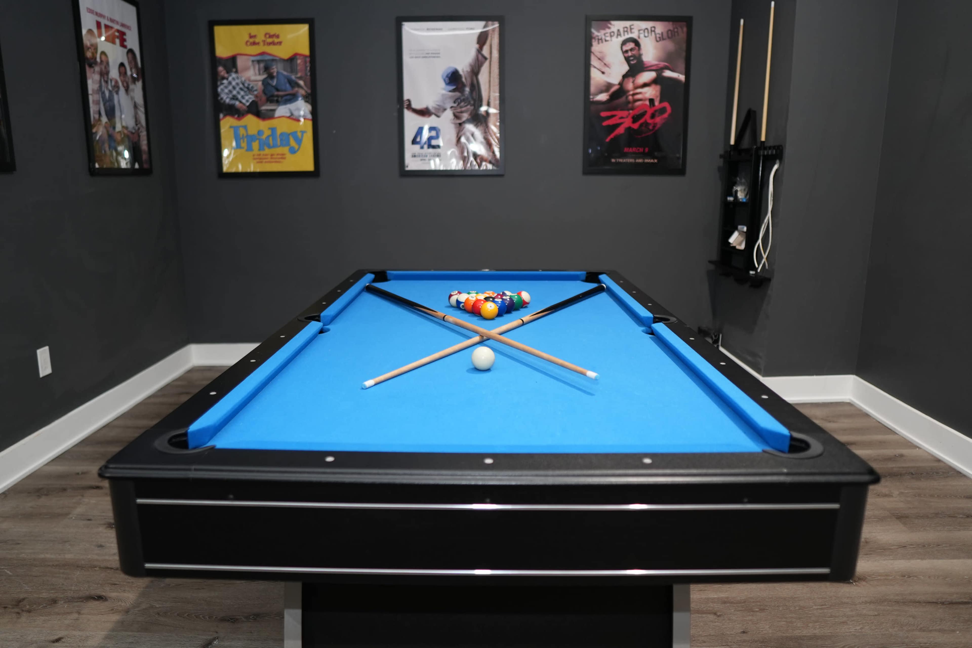 A blue pool table with four cue sticks crossed over a set of colored balls sits in a room decorated with framed movie posters on the walls.