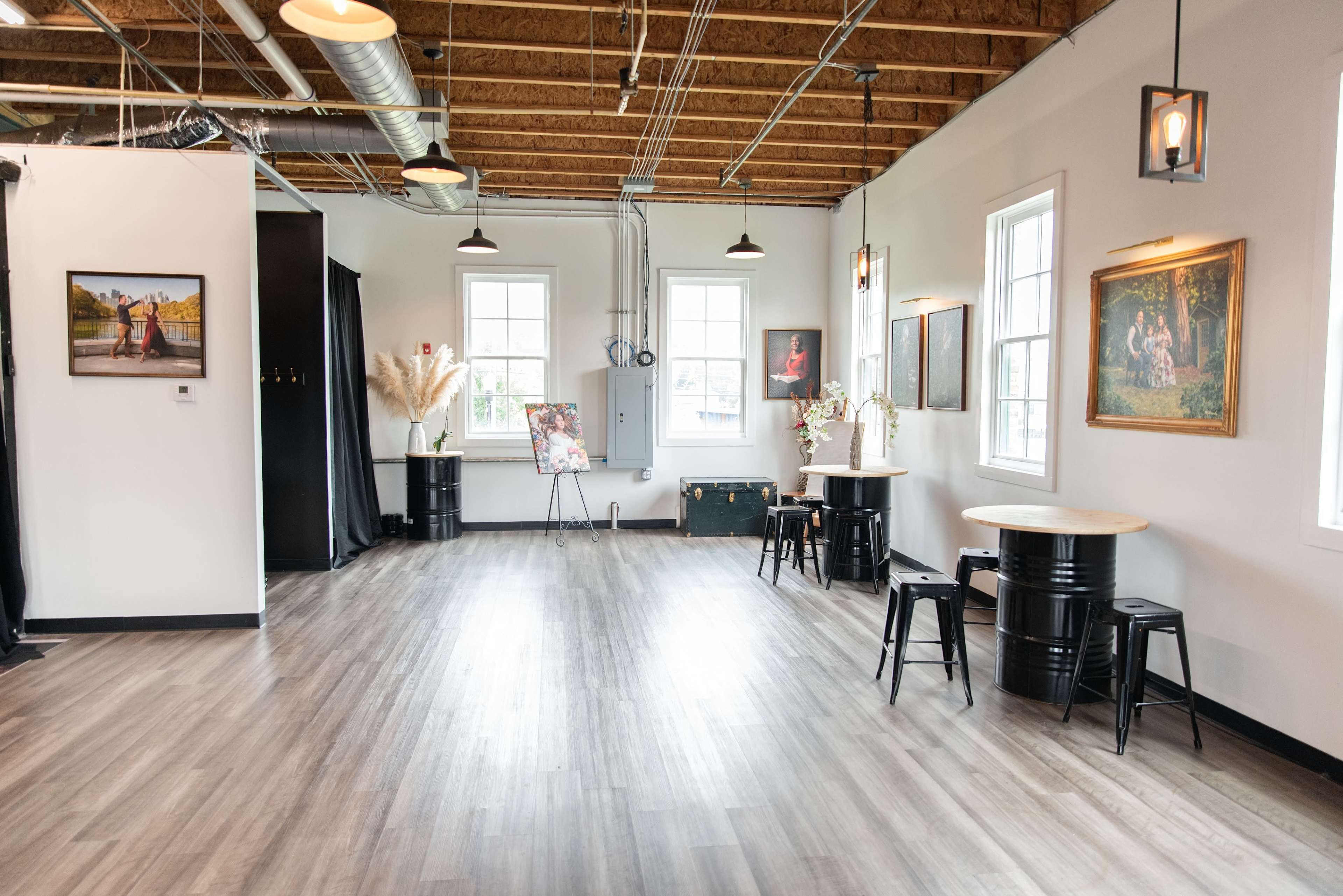 The image features a spacious, well-lit room with wooden flooring, showcasing several artworks on the walls and two round tables with black stools.