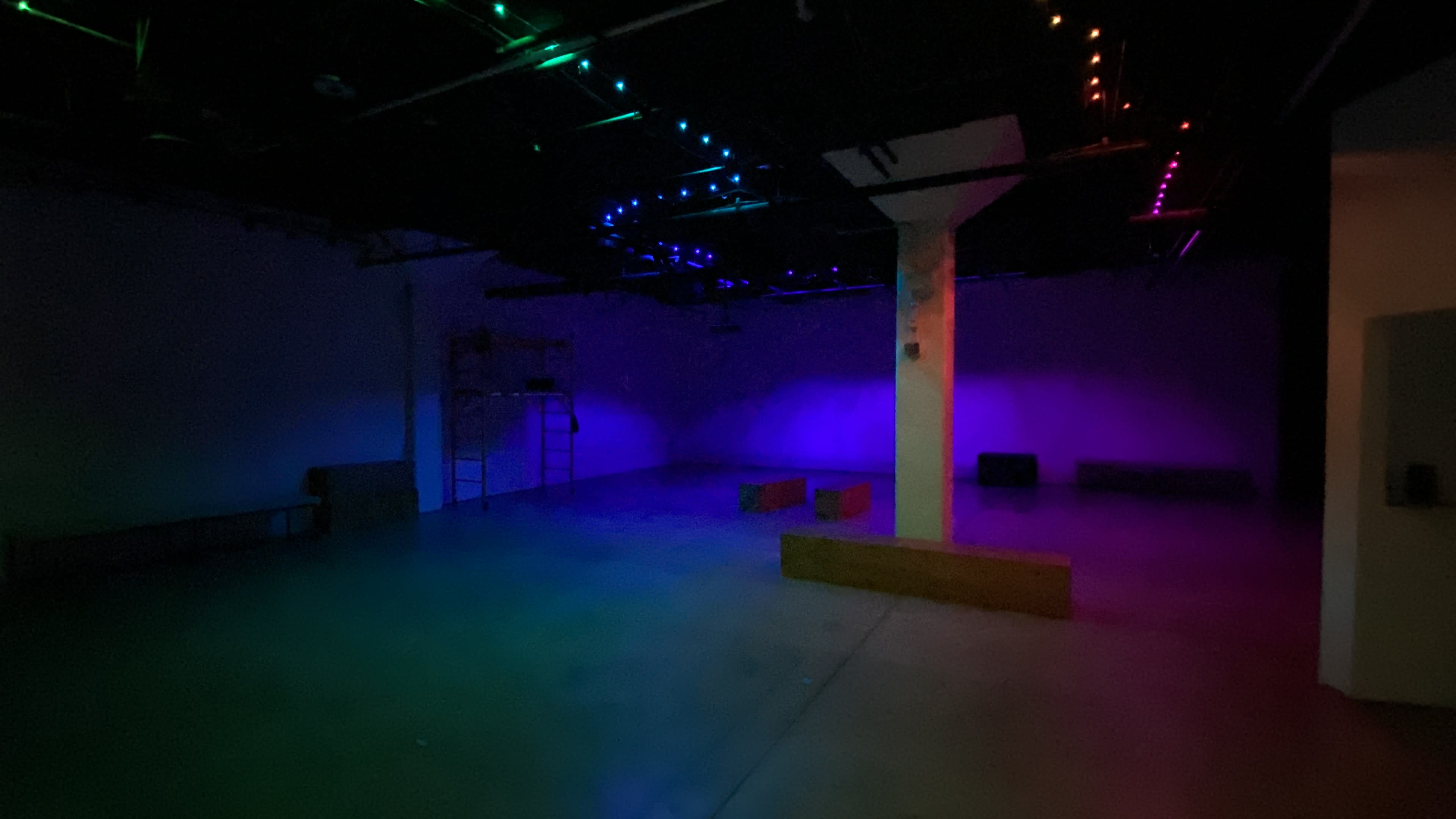 The image shows a dimly lit indoor space featuring a concrete floor, a central white column, and colorful LED lights along the ceiling.