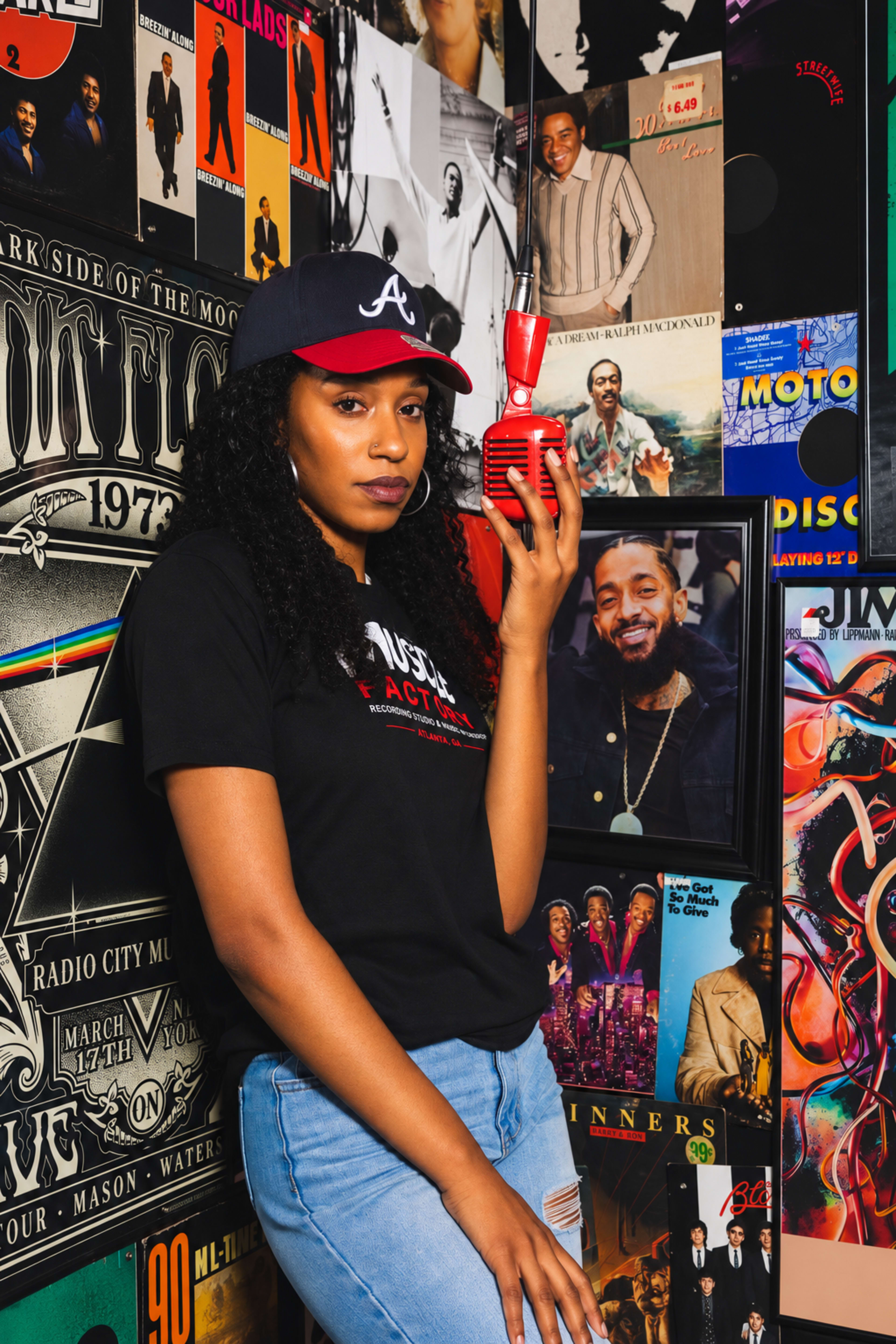 A woman stands next to a wall covered in music-themed posters, holding a red vintage microphone.