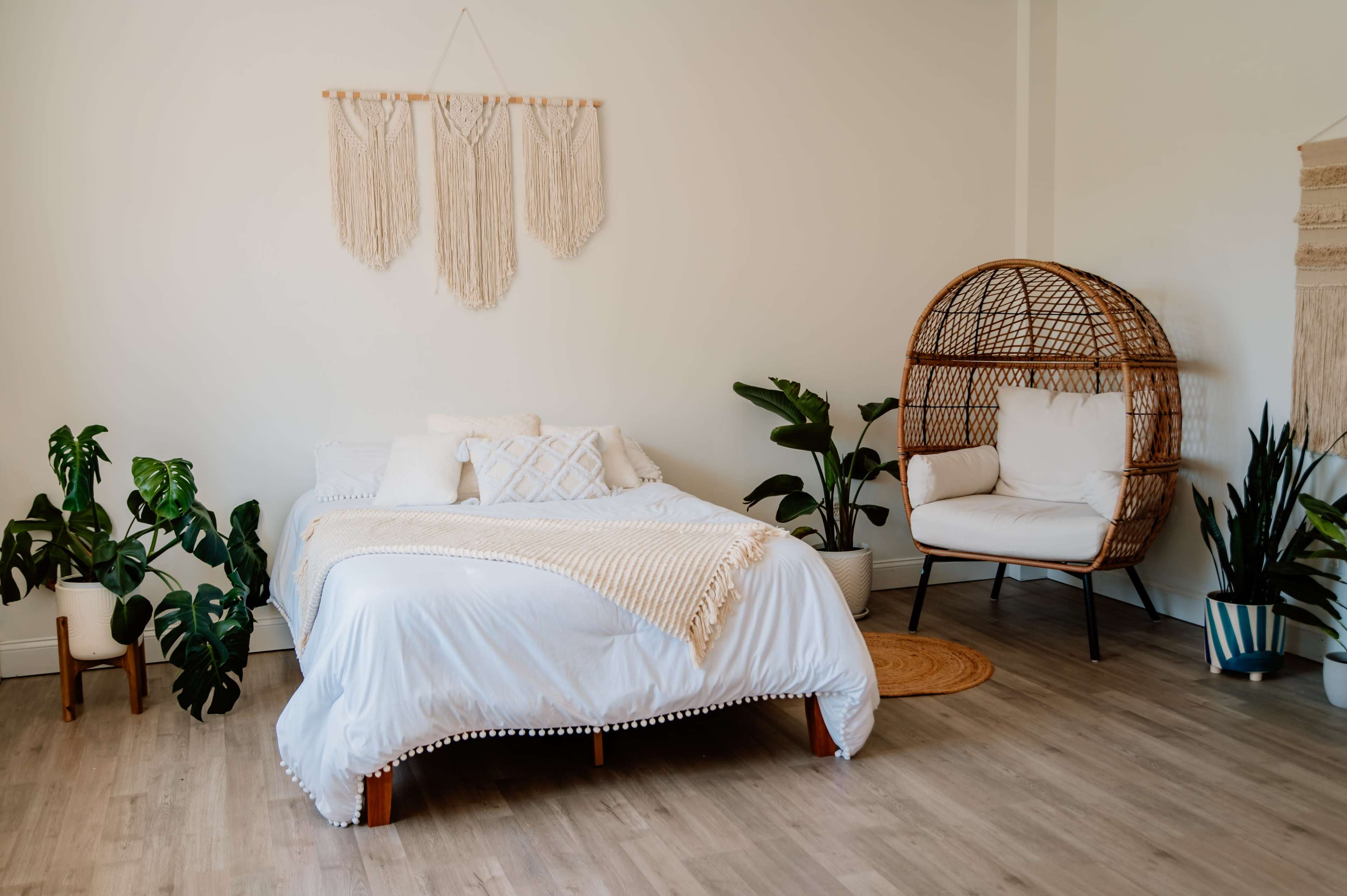 A neatly arranged bedroom with a white bed, decorative wall hangings, a cozy chair, and various indoor plants.