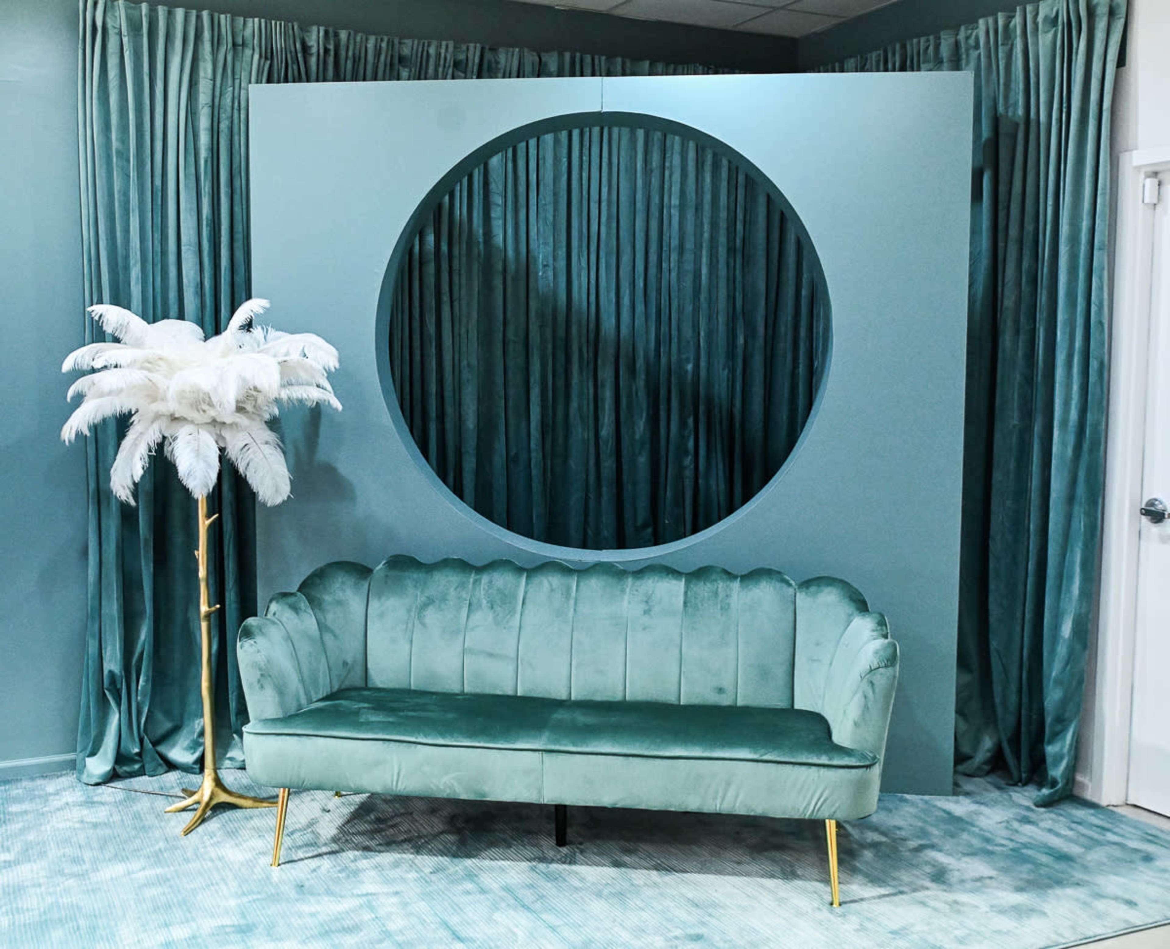 A green velvet sofa sits in front of a circular cut-out in a textured fabric backdrop, accompanied by a white feathered palm tree.