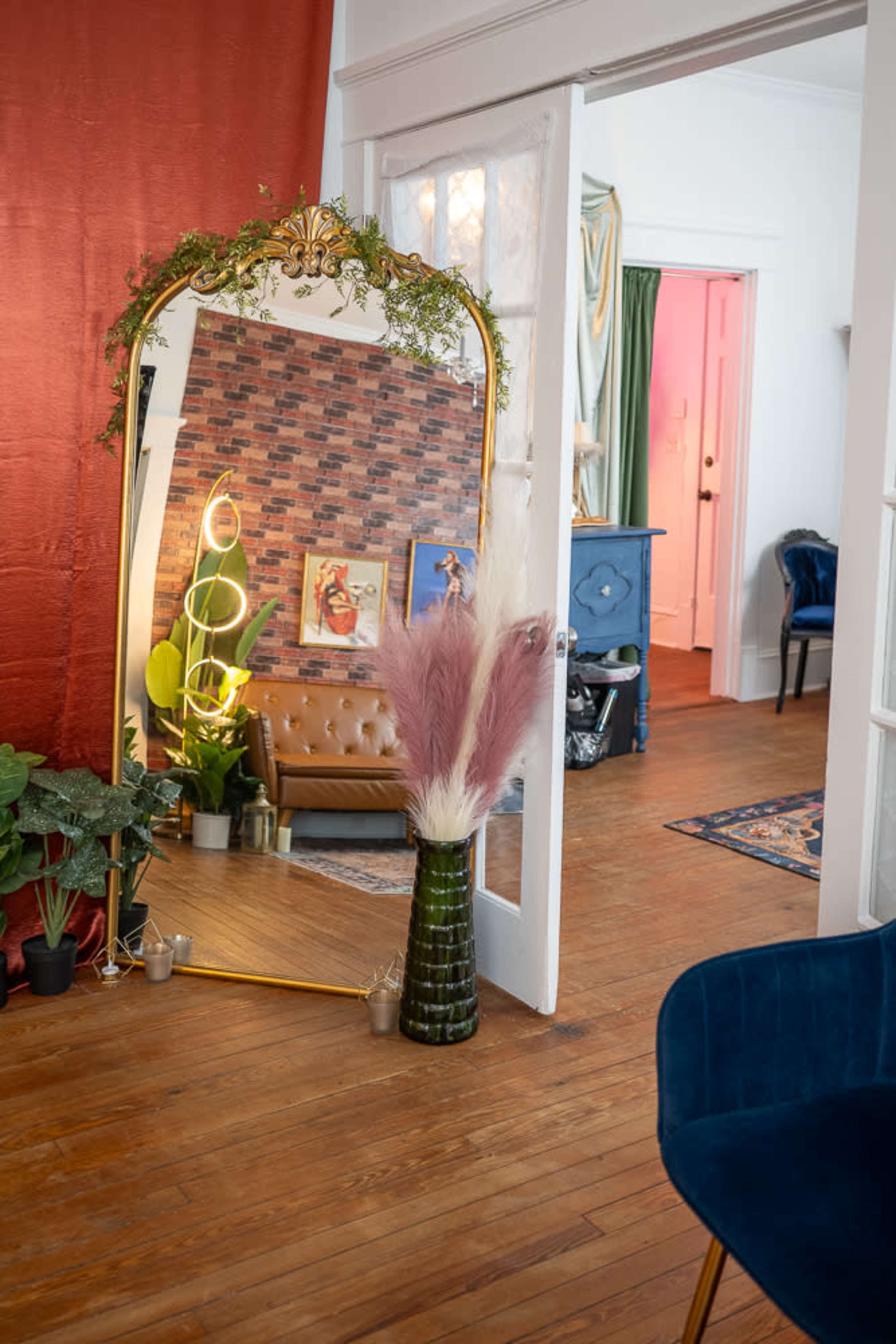 The image shows a decorated interior with a large mirror framed in gold, a vintage brown couch, potted plants, and a vibrant accent wall.