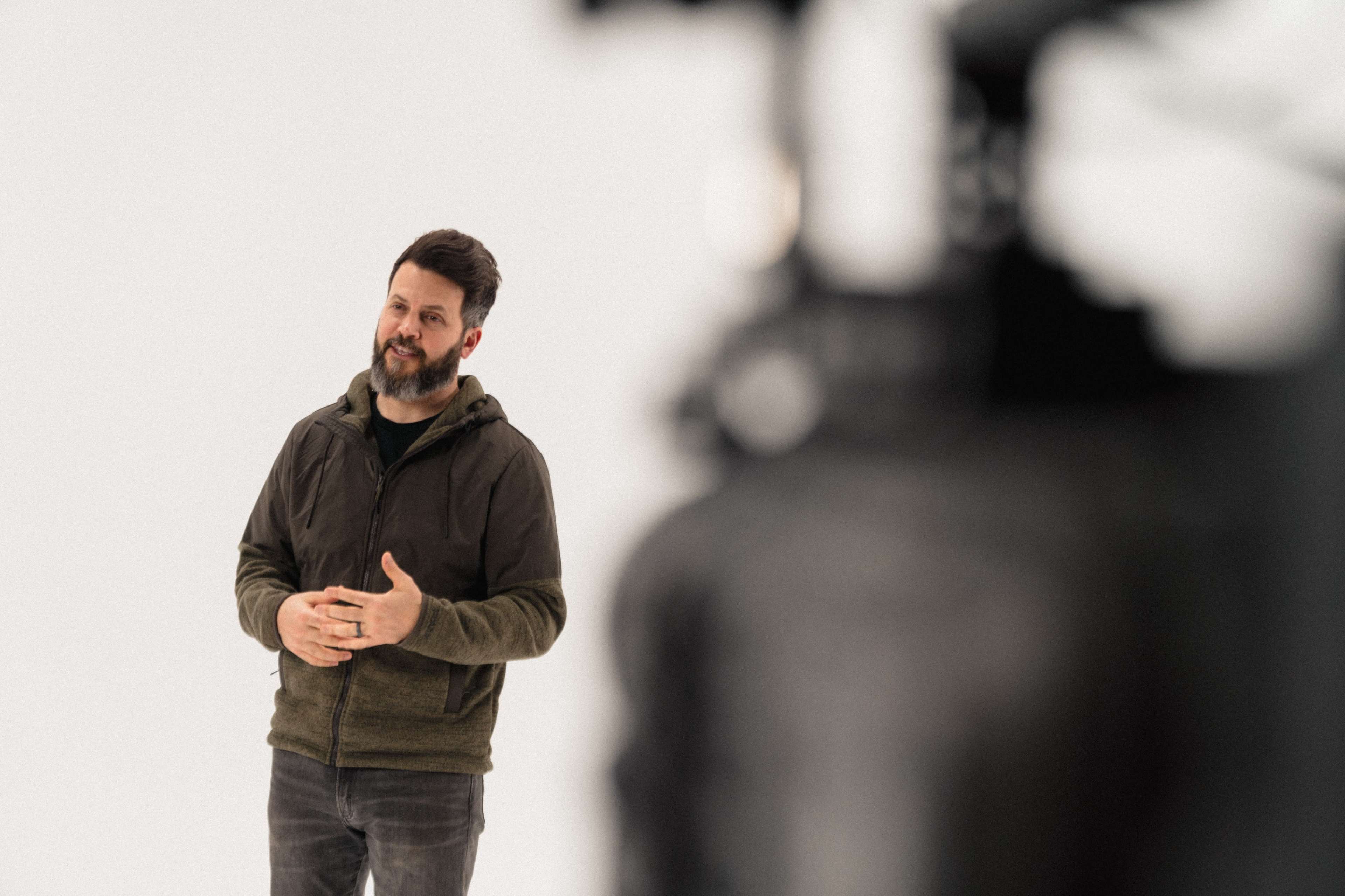 A man in a hooded jacket stands and speaks in front of a camera in a studio setting.