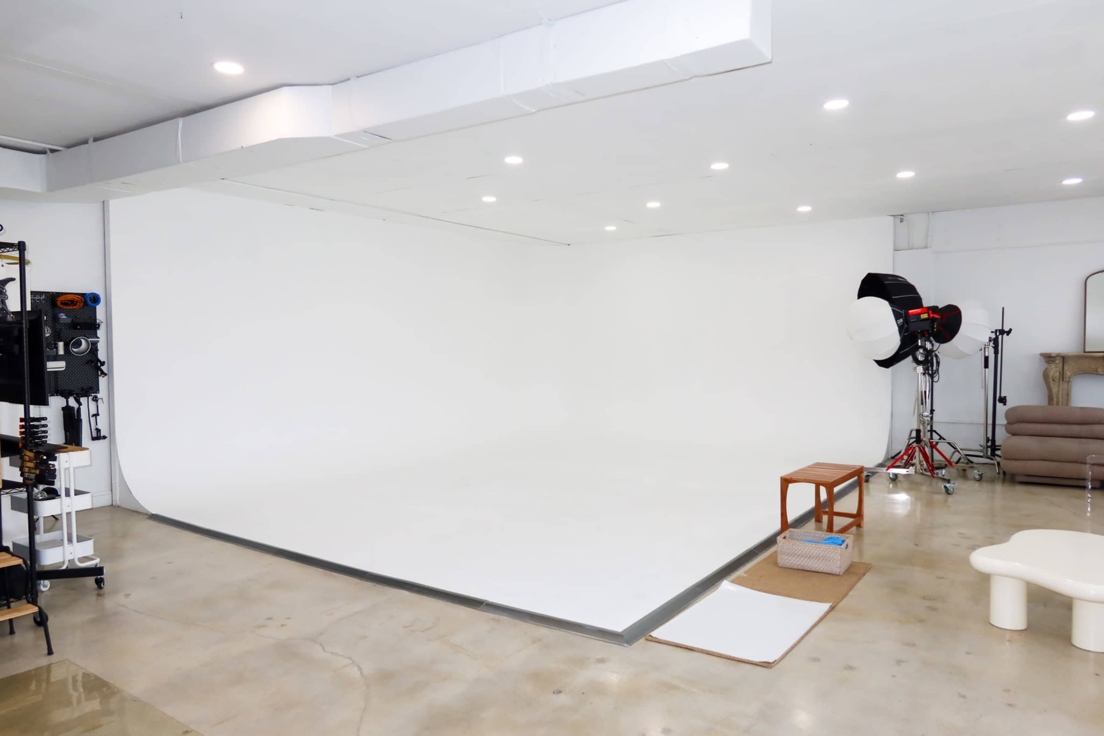 The image shows a photography studio with a large white backdrop, several lighting setups, and minimal furniture.