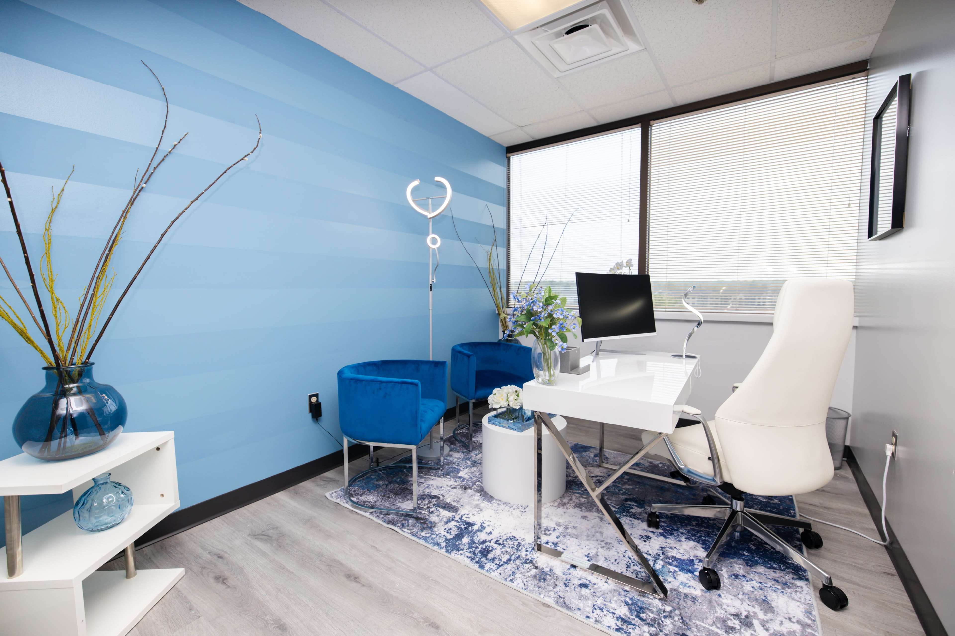 A modern office space featuring a white desk with a computer, two blue velvet chairs, and decorative plants in vases against a striped blue wall.