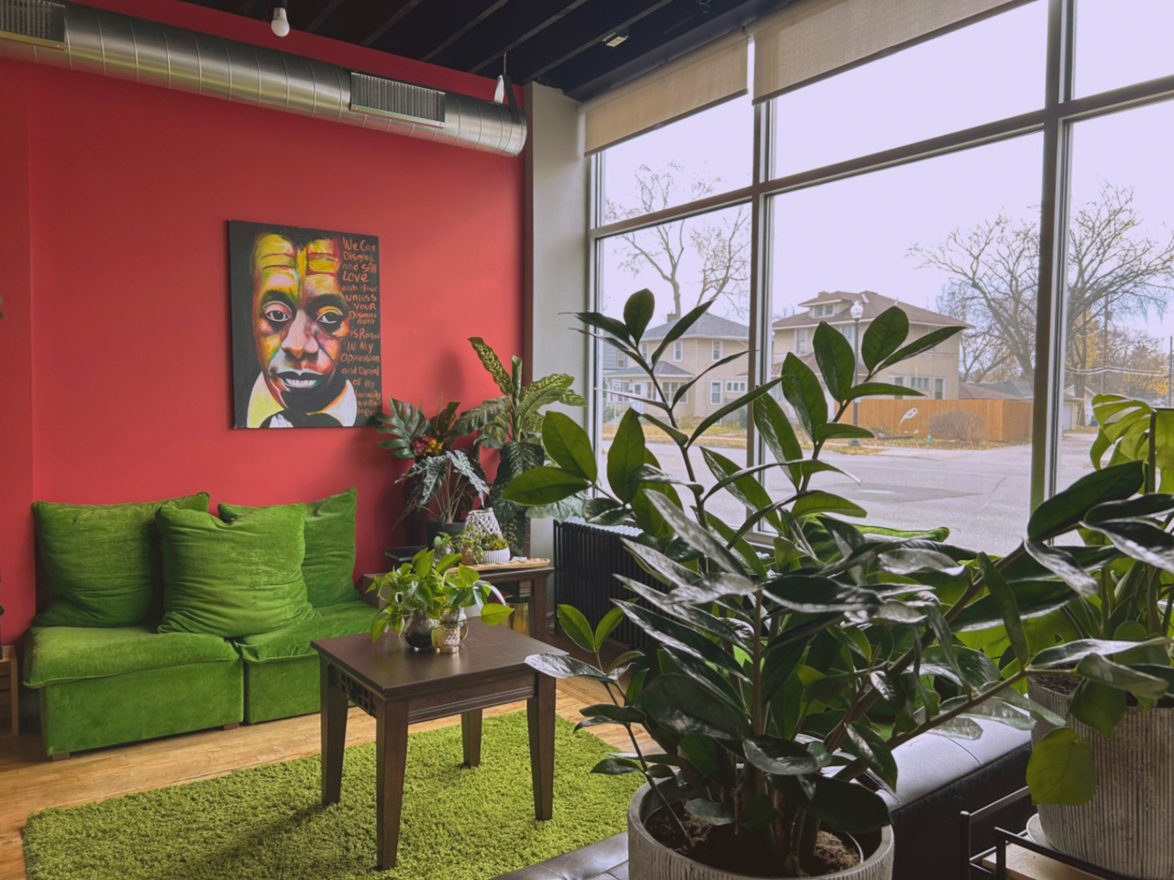 The scene features a cozy indoor space with green sofas, a table, and a large artwork hanging on a red wall, surrounded by several potted plants.