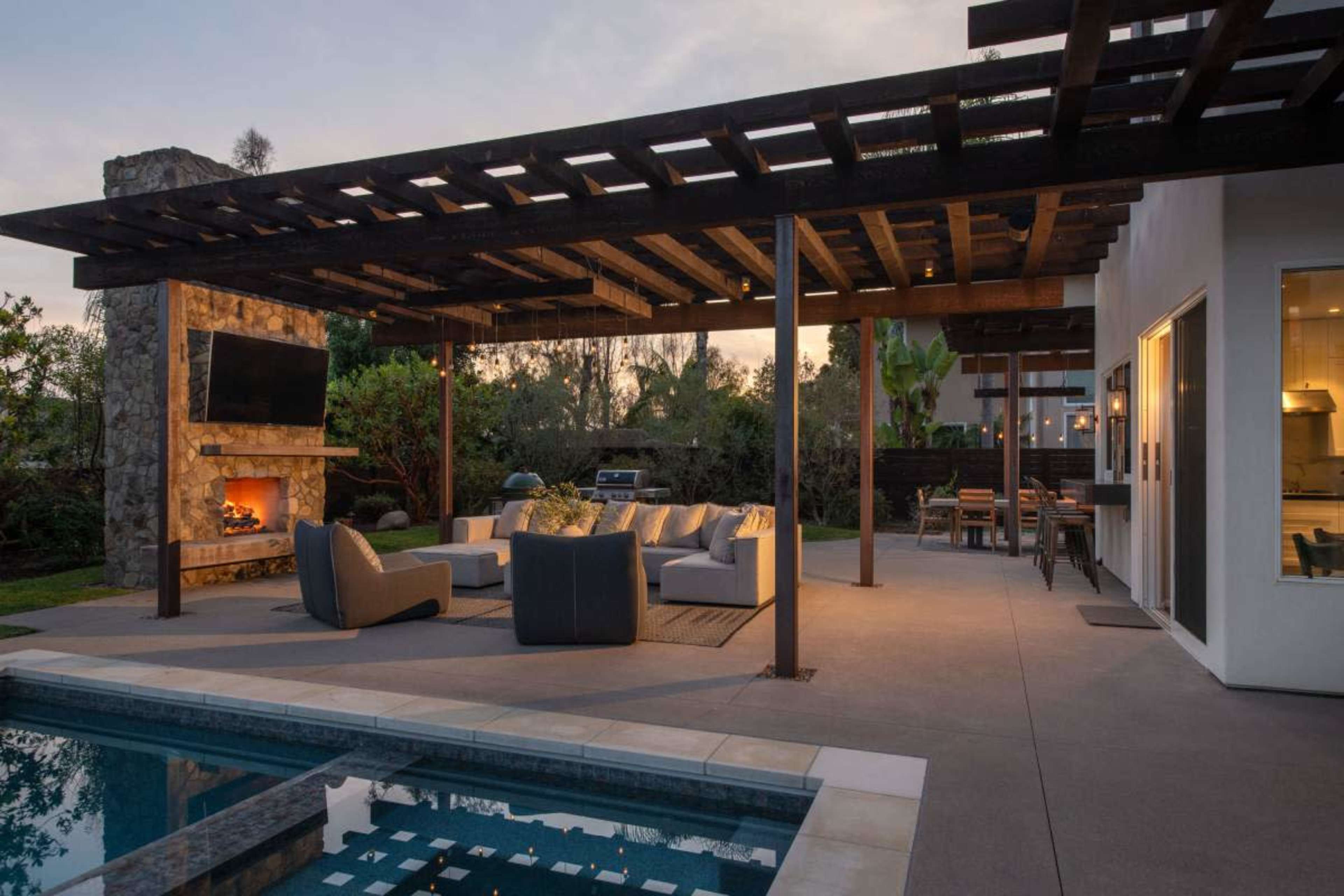 A modern outdoor living space features a pool, a stone fireplace, comfortable seating, and dining area under a wooden pergola.