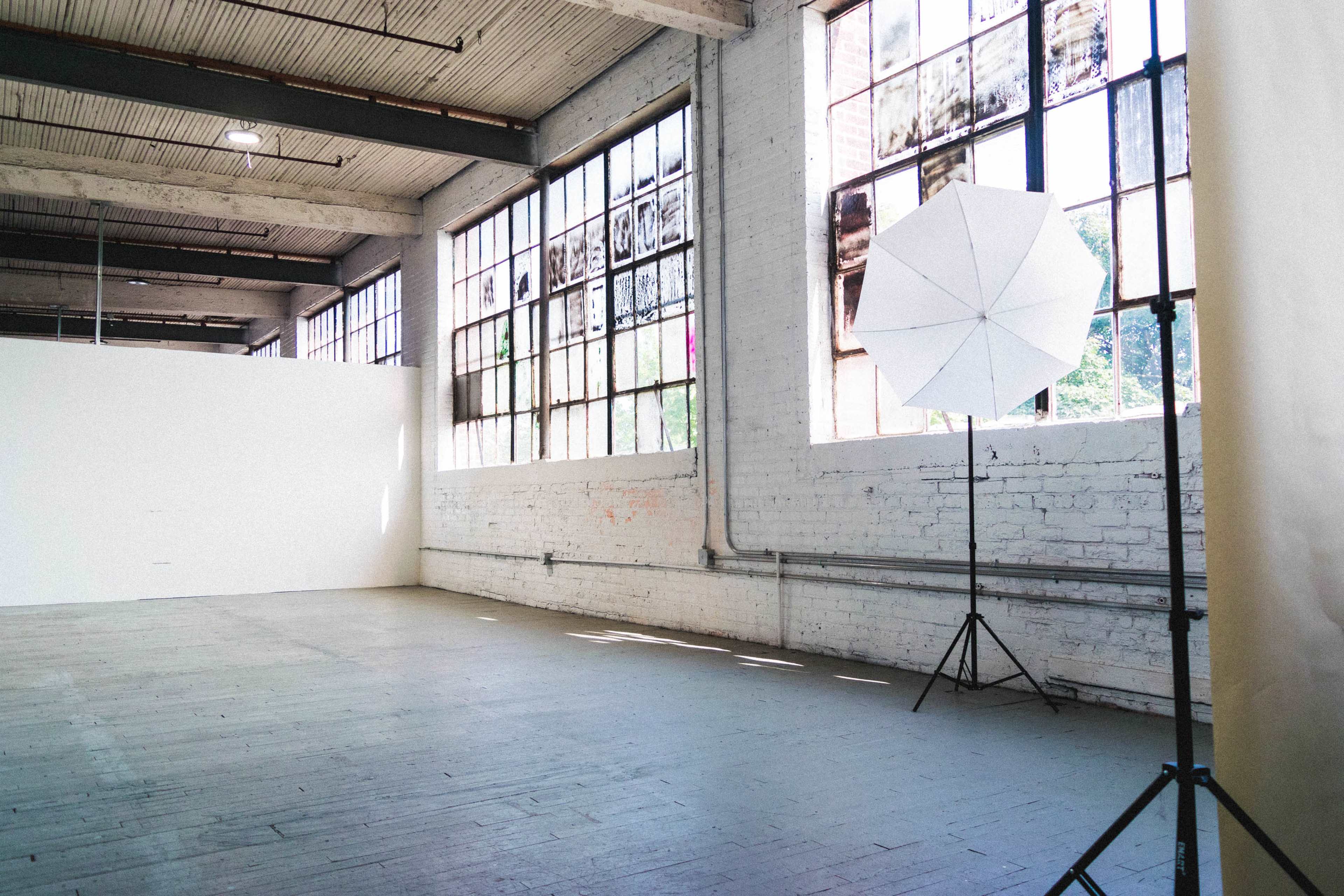 A spacious industrial room with large windows and a photography umbrella setup in one corner.