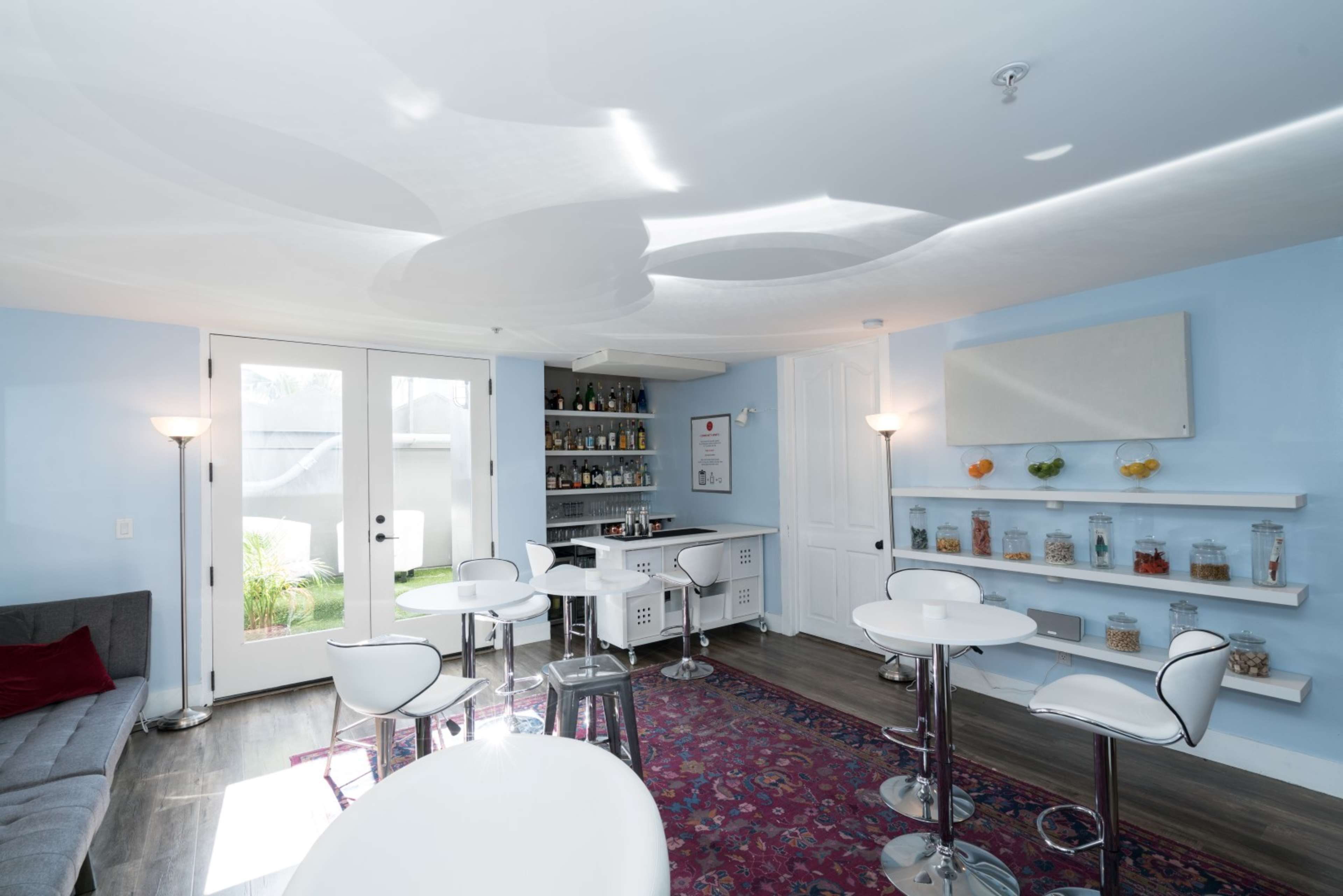 A modern lounge area with a bar and seating, featuring light blue walls, a patterned ceiling, and glass jars on shelves.