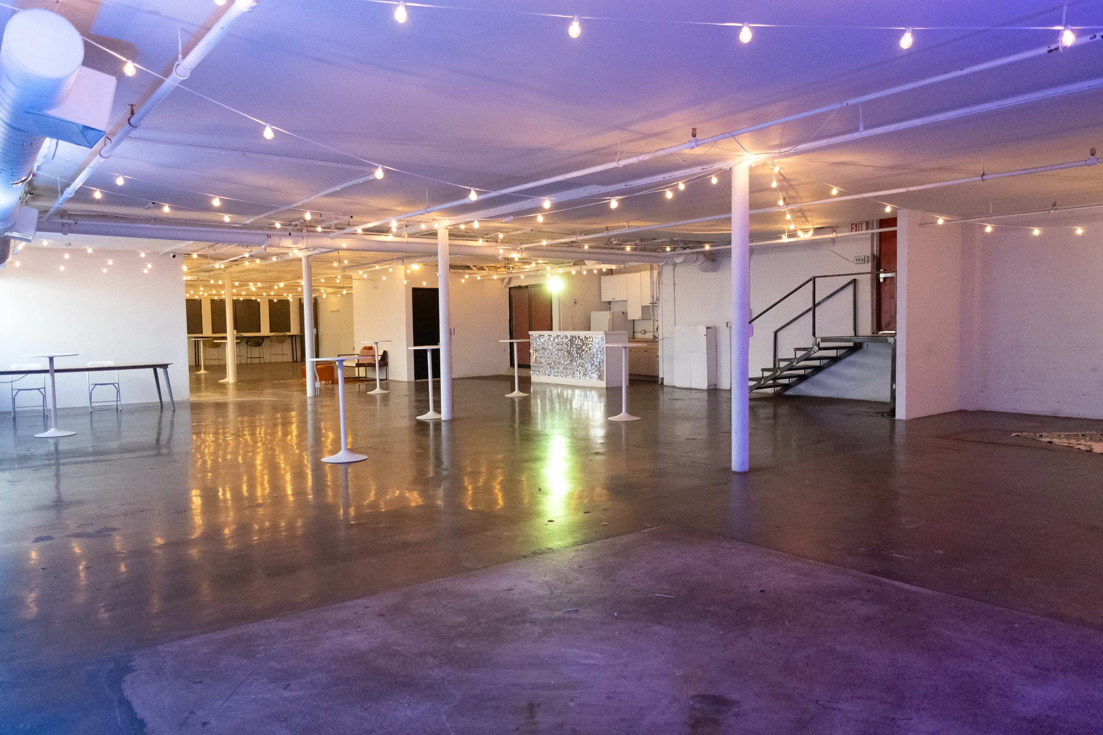 The image shows a spacious, softly lit event venue with string lights overhead, featuring bare concrete floors and several standing tables.