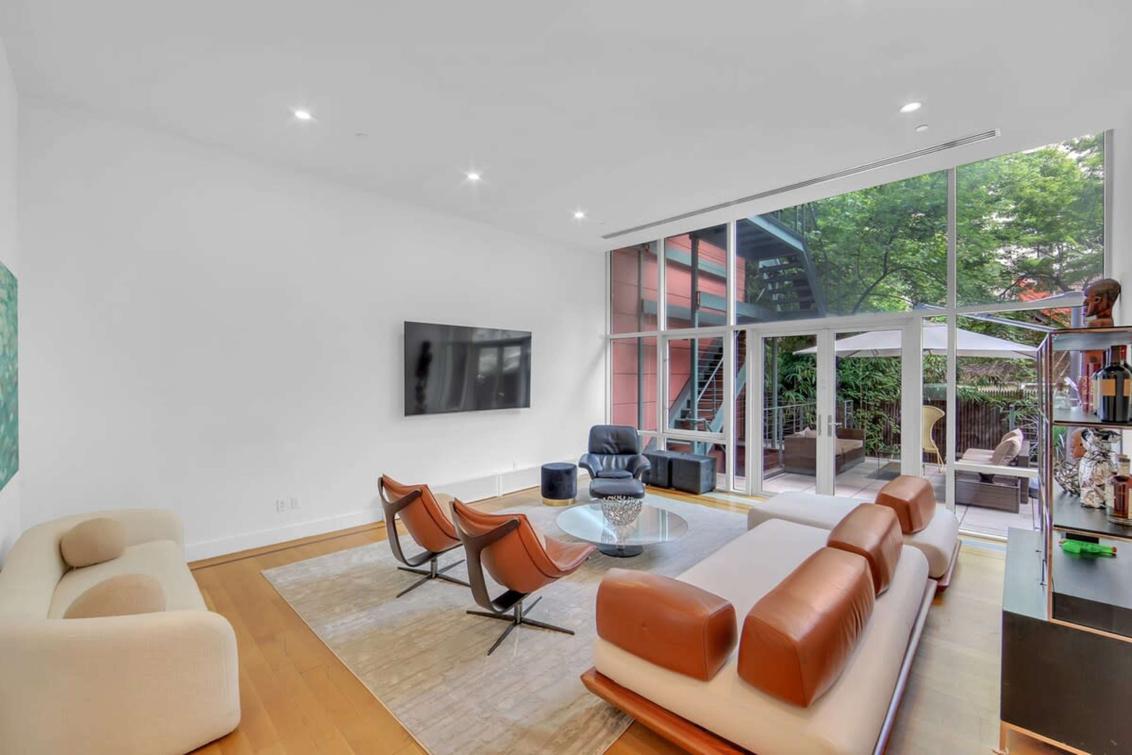The image shows a modern living room featuring a large television, contemporary seating, a round glass coffee table, and large windows that open to an outdoor space.