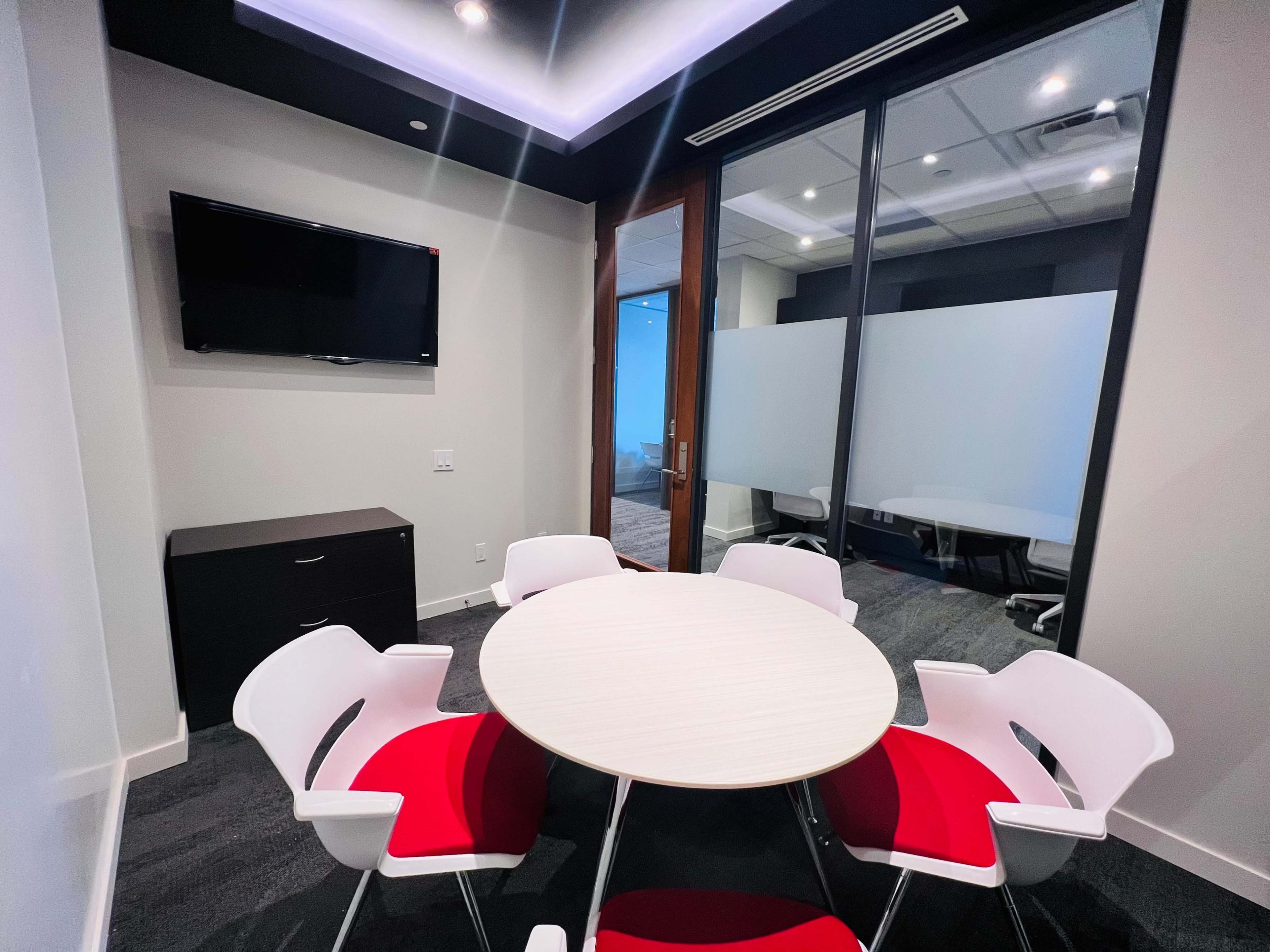 A small conference room features a round table with white and red chairs, a flat-screen TV on the wall, and glass partitions.
