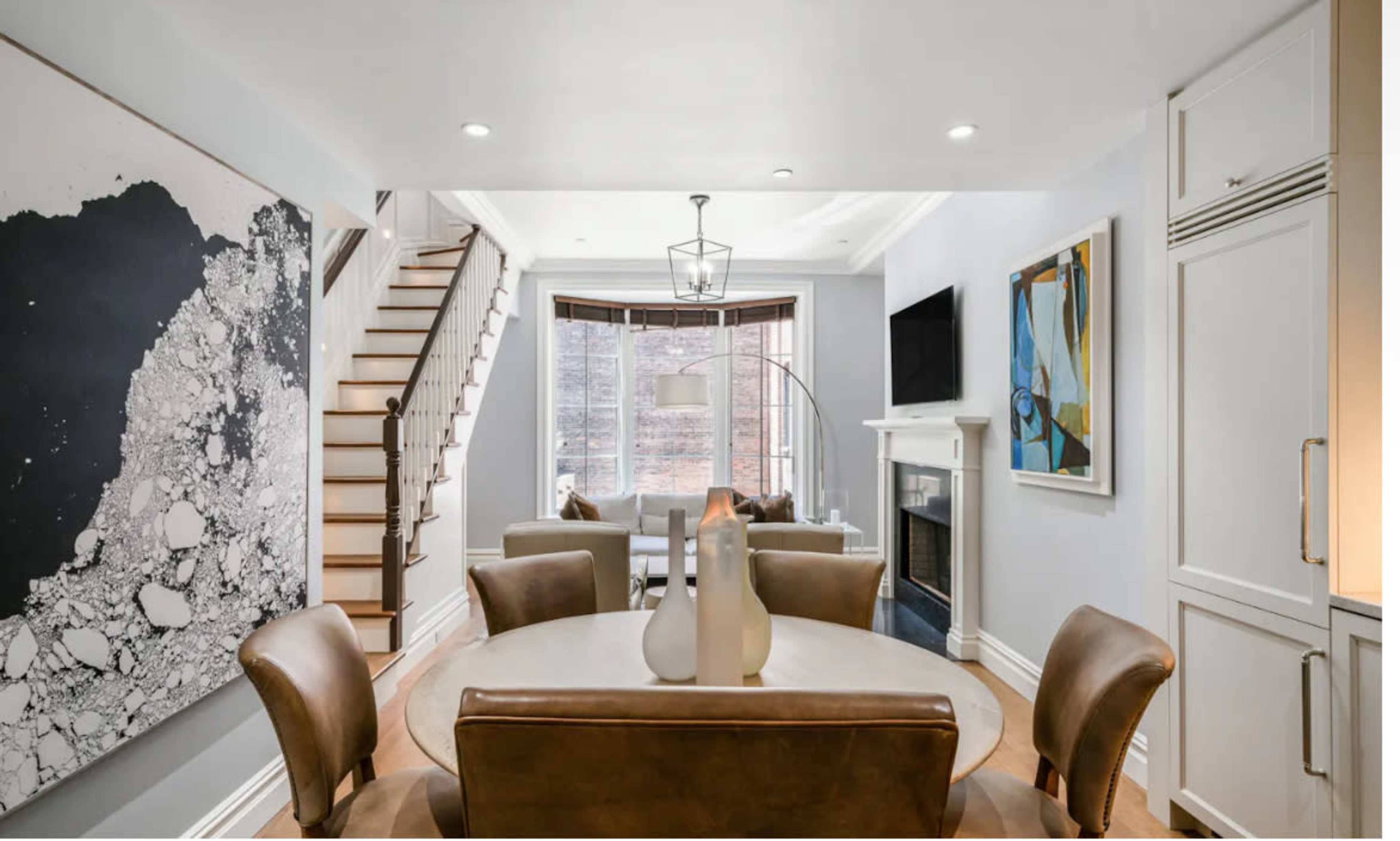 The image shows a modern dining area with a circular table and leather chairs, adjacent to a staircase and a living space featuring artwork and a fireplace.
