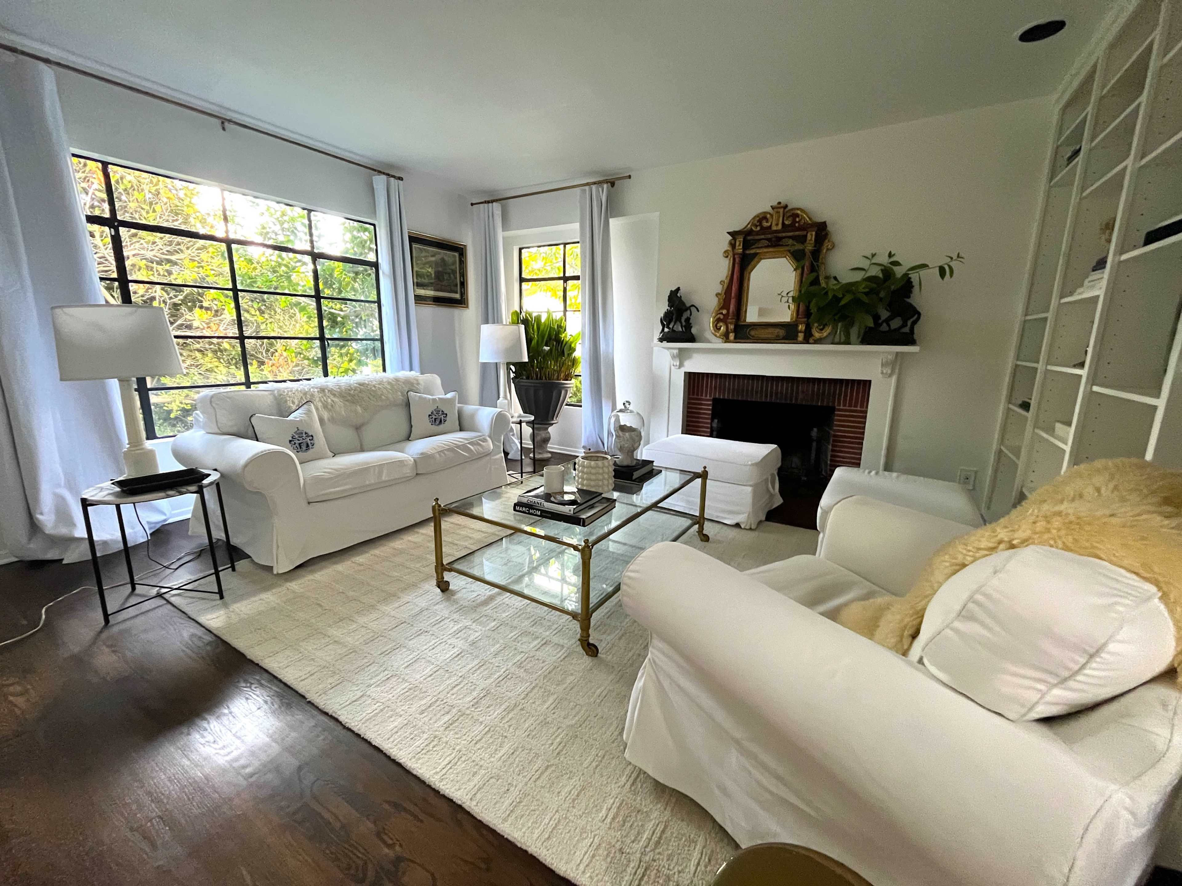 The living room features white sofas, a glass coffee table, and a fireplace surrounded by decorative plants and artwork, with large windows allowing natural light to fill the space.
