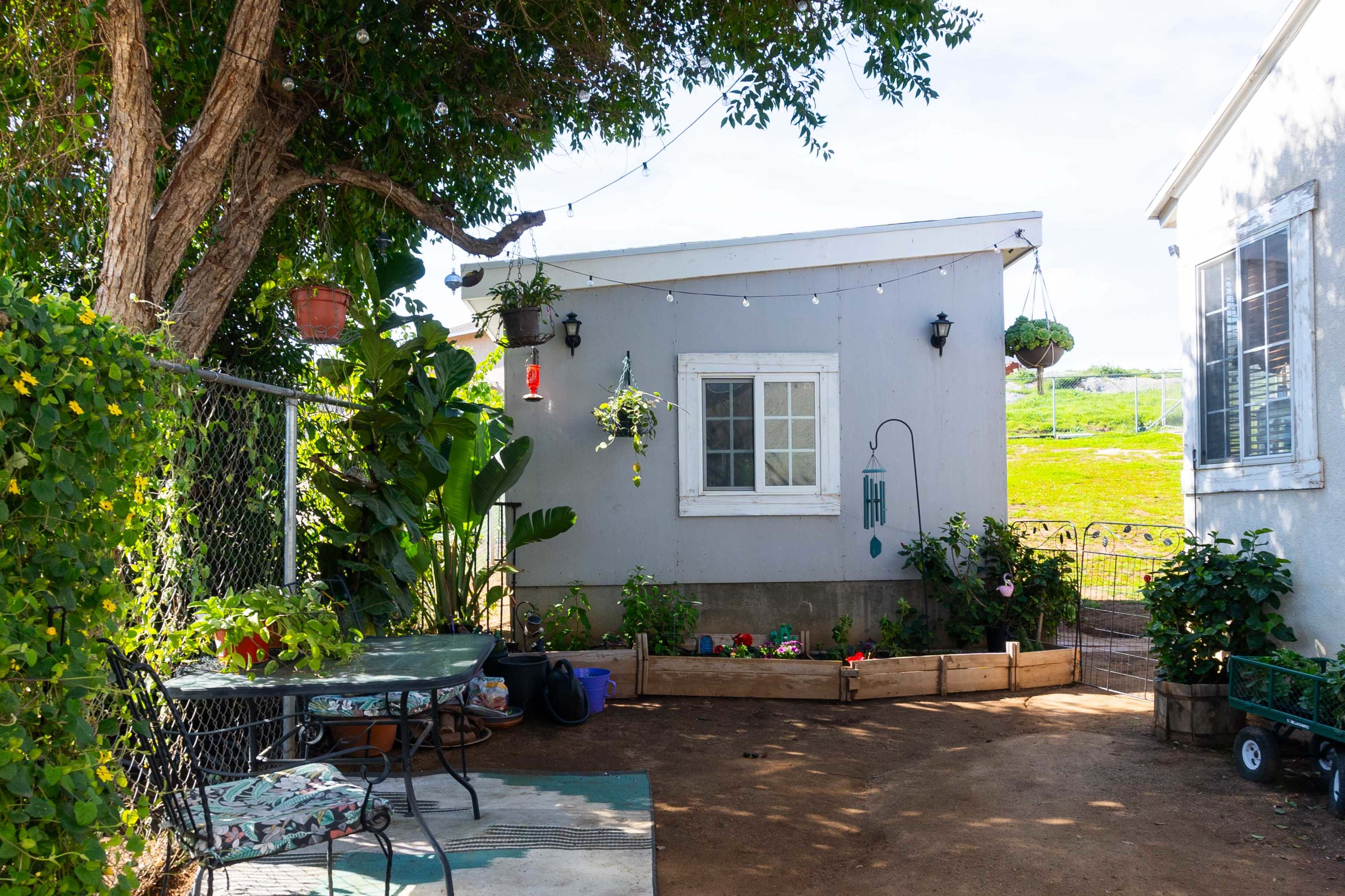 A small gray house with a white window is surrounded by greenery and features a seating area with a table and chairs.