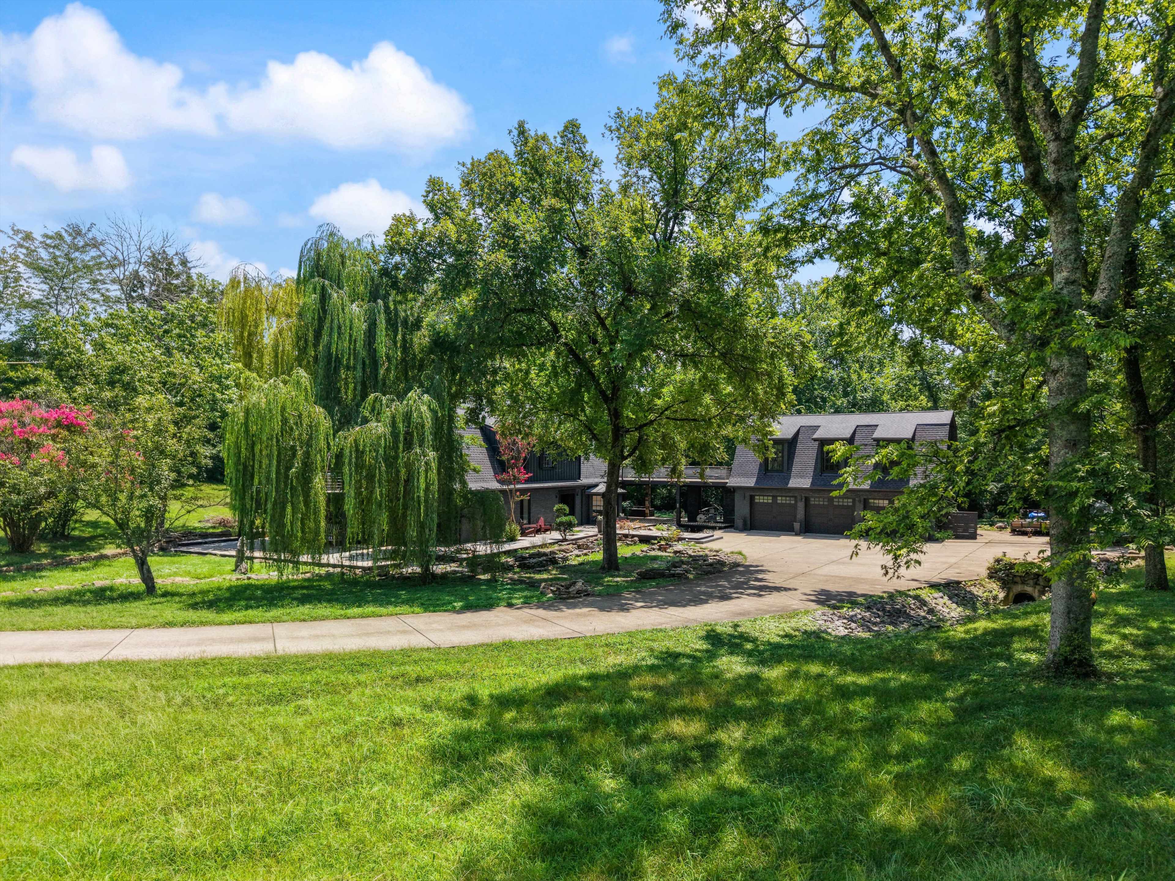 The image shows a large house nestled among trees, with a curved driveway leading to the property and a weeping willow in the foreground.