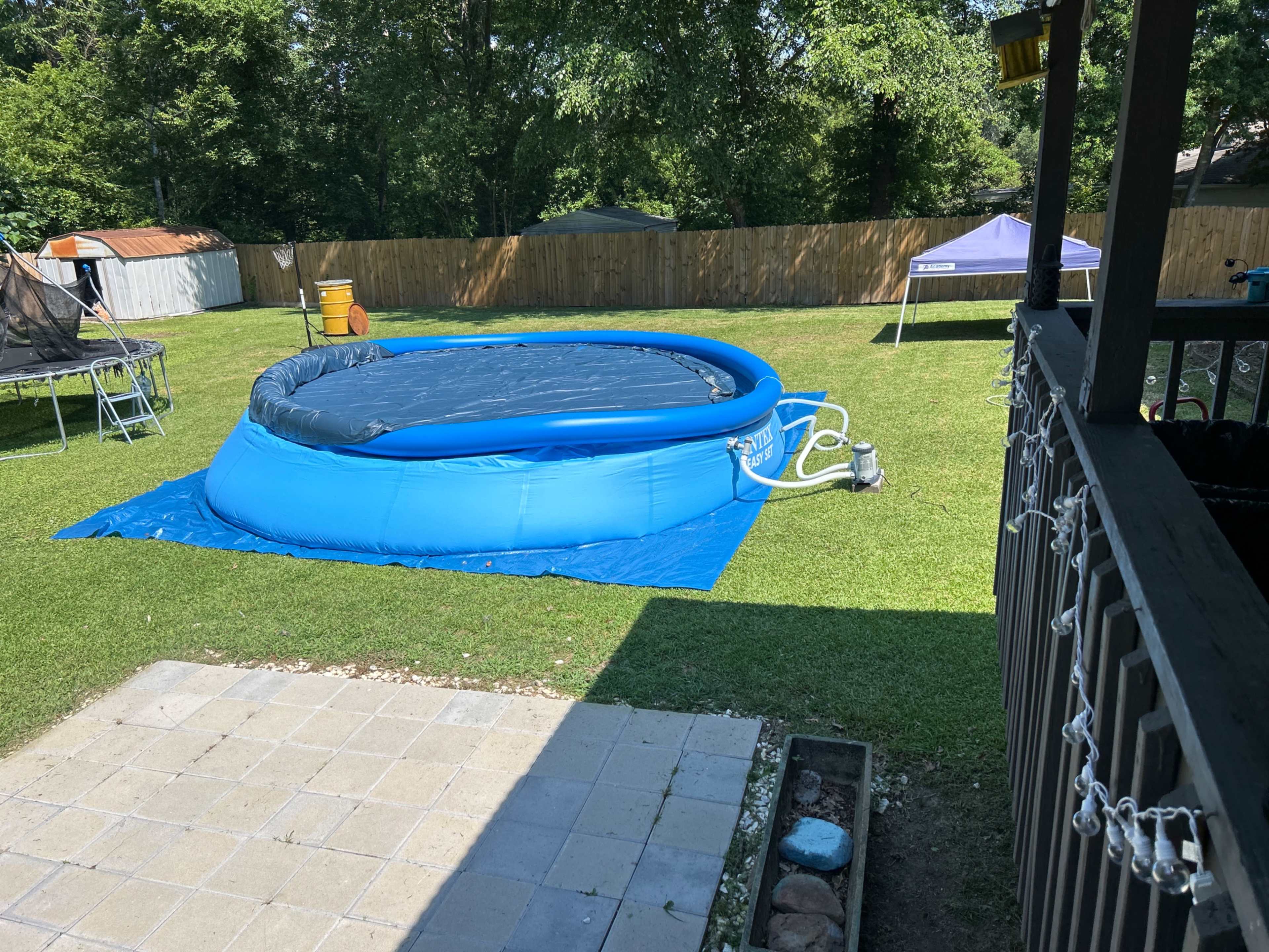 A blue inflatable pool sits on a grassy yard surrounded by trees and a wooden fence.