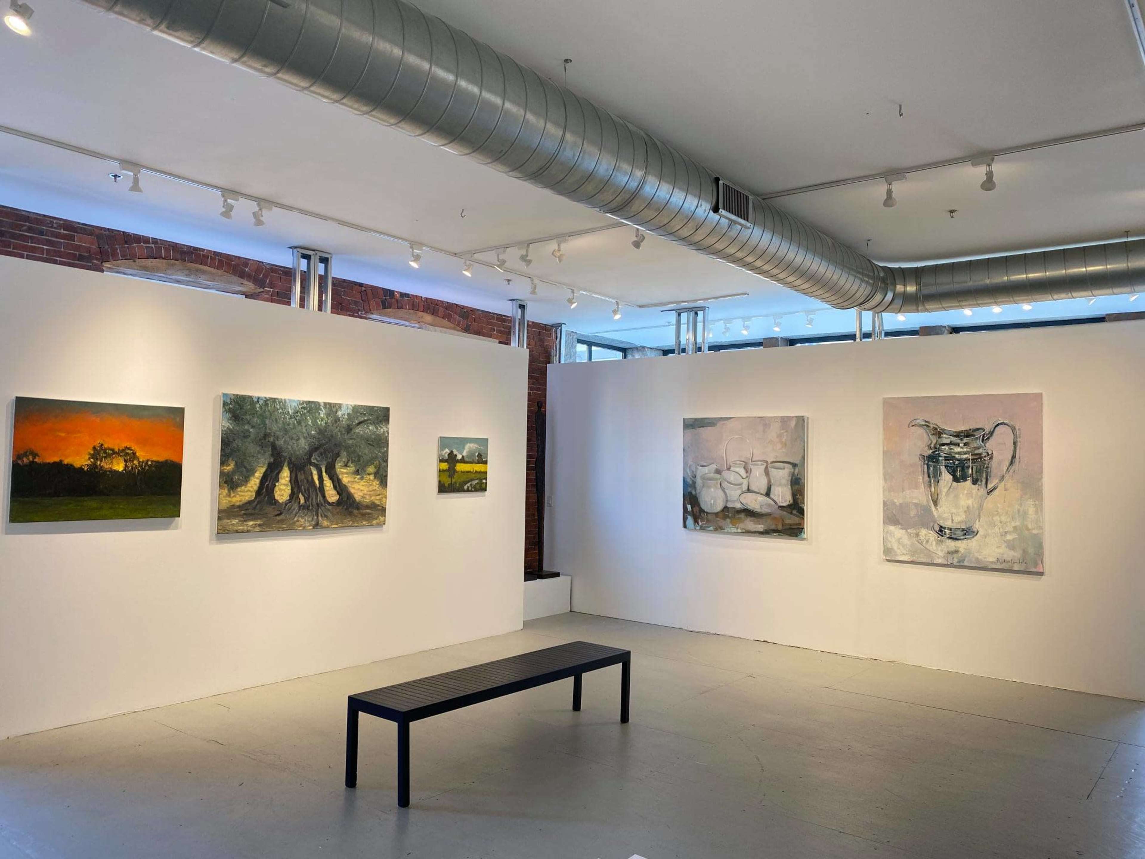 An art gallery with several paintings displayed on white walls, a bench in the center, and exposed ceiling ductwork visible above.