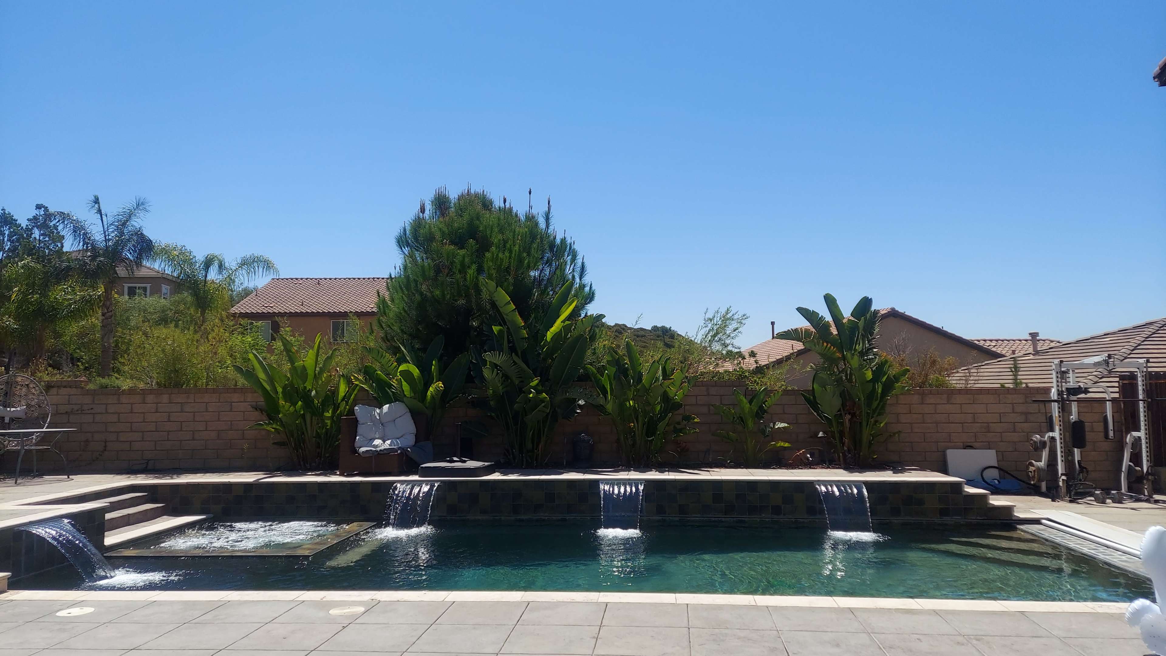 The image shows a swimming pool with cascading water features, surrounded by tall plants and houses under a clear blue sky.